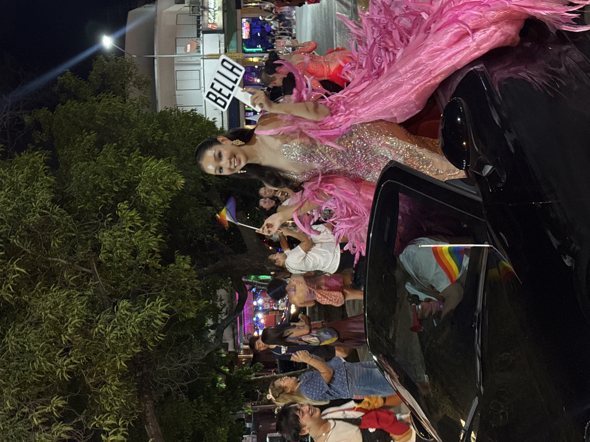 The image captures a nighttime scene in Chiang Mai City Municipality, Thailand. A woman is standing on a car. She wears a sparkly dress with a pink feathered collar and holds a sign that says "BELLA" and a rainbow flag. The car seems to be part of a parade, with other people visible in the background. Some hold rainbow flags. The setting is a street with trees and buildings, with lights illuminating the scene.
