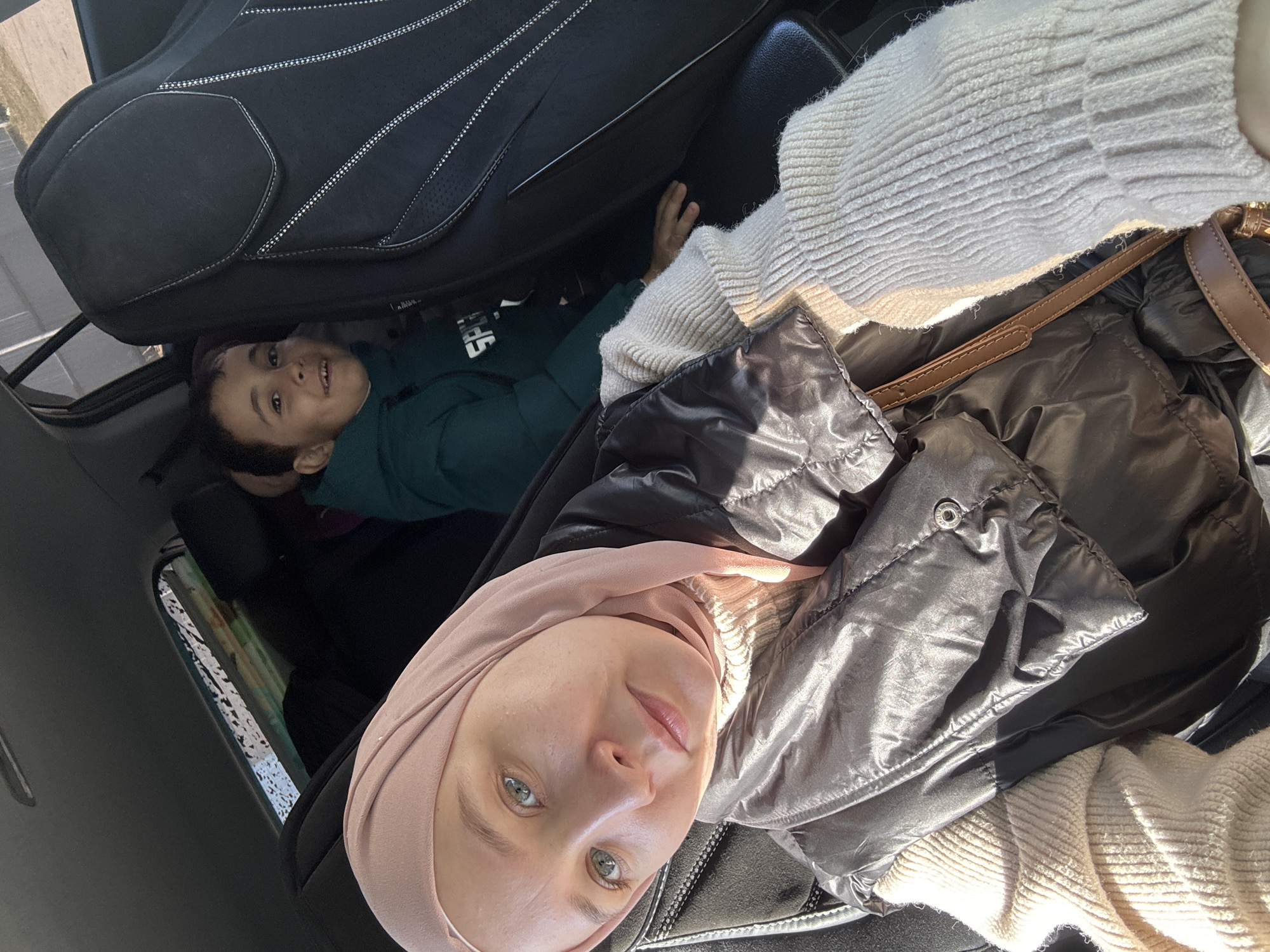 The image shows a woman wearing a pink hijab, a cream-colored sweater, and a dark puffer jacket, possibly taking a selfie inside a car. Next to her, in the back of the car, a child wearing a green shirt is visible. A car seat is also present. The location is not discernible from the visual information provided.