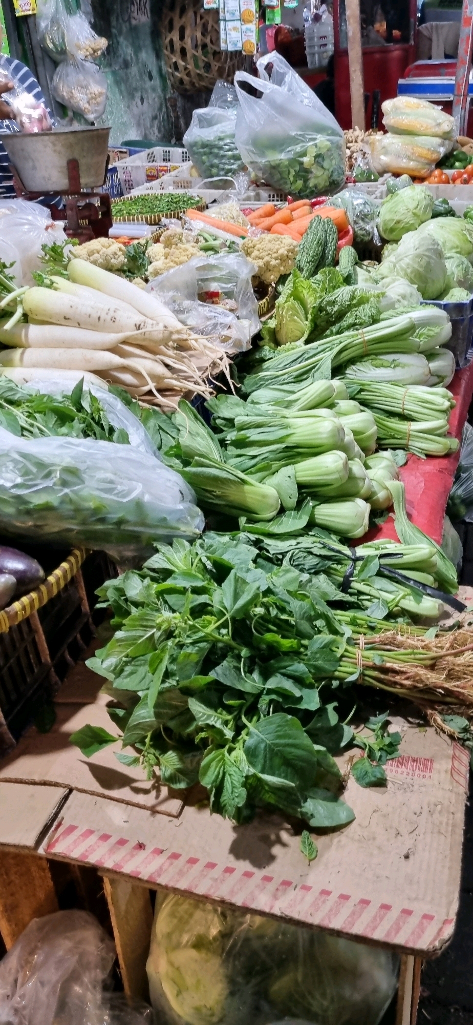 In Denpasar, Indonesia, this image captures a vibrant scene at a market stall laden with fresh produce. A variety of vegetables, including bundles of leafy greens such as bok choy and spinach, are prominently displayed. White radishes are stacked neatly, alongside cauliflower florets and orange carrots. Other vegetables like cabbage and corn are also visible, some wrapped in plastic. The stall features woven baskets and plastic bags, indicating a typical market setting. The overall impression is one of abundance and freshness.