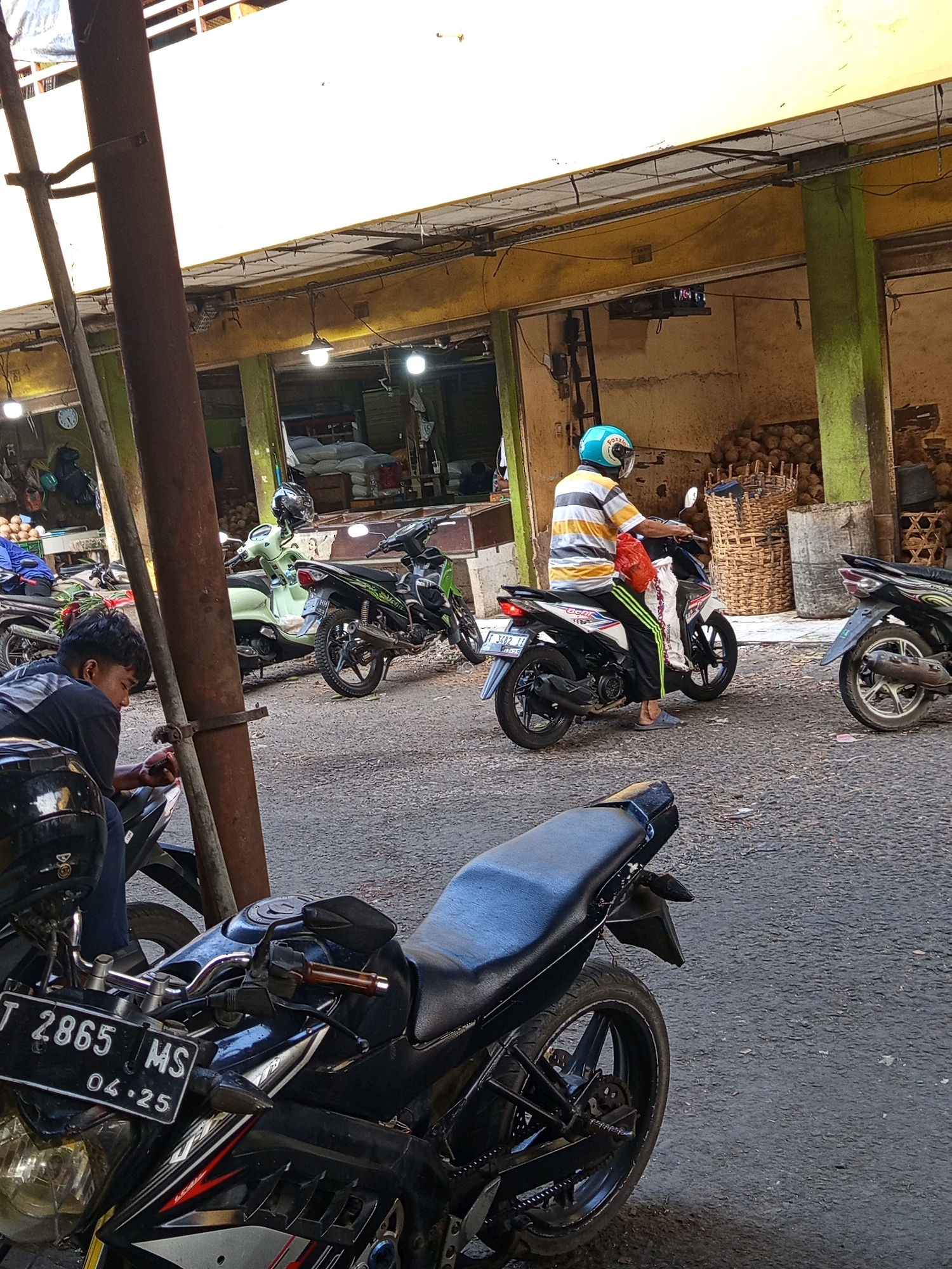The image captures a street scene in what appears to be a market area in Karawang, Indonesia, under bright daylight. The foreground is dominated by the back portion of a black motorcycle, with its license plate visible, reading "T 2865 MS 04.25". To the left, a young man wearing a black t-shirt sits on a motorcycle, looking down at his phone. He is partially obscured by a thick, dark brown vertical pole.
In the mid-ground, a person wearing a striped t-shirt and a blue helmet rides a white and black motorcycle. They are carrying a red bag and a white bag on the motorcycle. Behind them, several other motorcycles are parked, including a light green one. The background reveals the facade of market stalls or buildings with yellow and green architectural elements. Large woven baskets, possibly containing produce like coconuts, are stacked to the right. The ground is paved with asphalt.