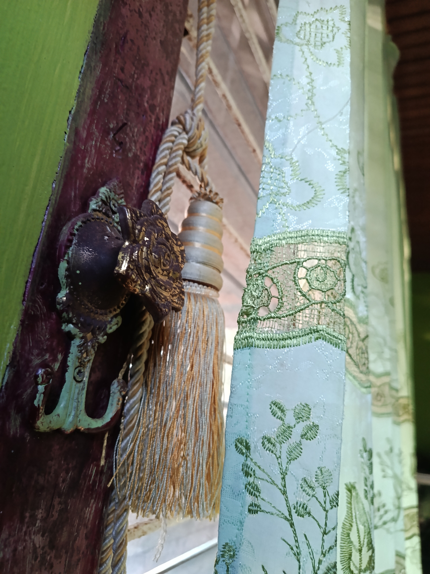 An ornate, antique-style curtain tie-back hook is attached to a dark red, weathered wooden beam, which is painted green on its left side. The hook is made of a dark, possibly bronze or aged brass material, with intricate floral detailing, and shows signs of wear and some green patina. A thick, braided cord, cream and gold in color, is looped through the hook and extends downwards, supporting two decorative tassels. These tassels are also made of cream and gold threads, with cylindrical, metallic elements interspersed within them. To the right of the hook and tassels, a light blue curtain with a delicate, embroidered floral pattern in green is visible. The curtain has a lace-like border with a repeating circular motif. The background suggests an indoor setting with a window, indicated by the presence of wooden blinds behind the curtain and hints of natural light. The scene appears to be located in Mansalean, Indonesia, implying a residential or possibly a historic building context.