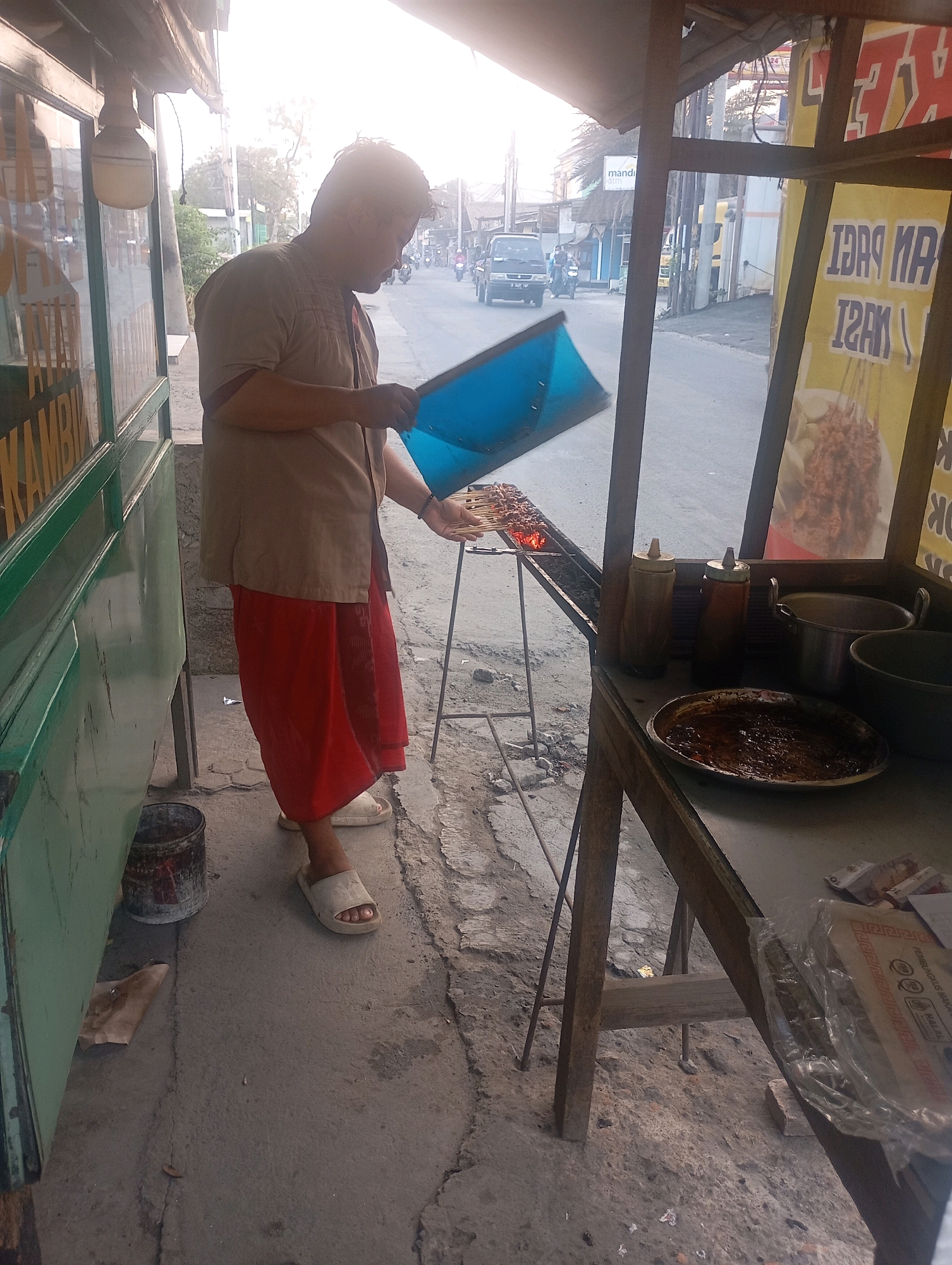 A man is shown grilling satay in front of a food stall on the side of a street in Babelan, Indonesia. The man, wearing a beige shirt and a red sarong, is holding a blue fan to stoke the charcoal grill. The stall appears to be constructed from wood and metal, with a green painted cabinet on the left side. On a wooden table to the right, there are bottles of sauce, a metal pot, and a plate with satay sauce. In the background, a street is visible with vehicles and buildings. The overall scene suggests a typical street food vendor operation.