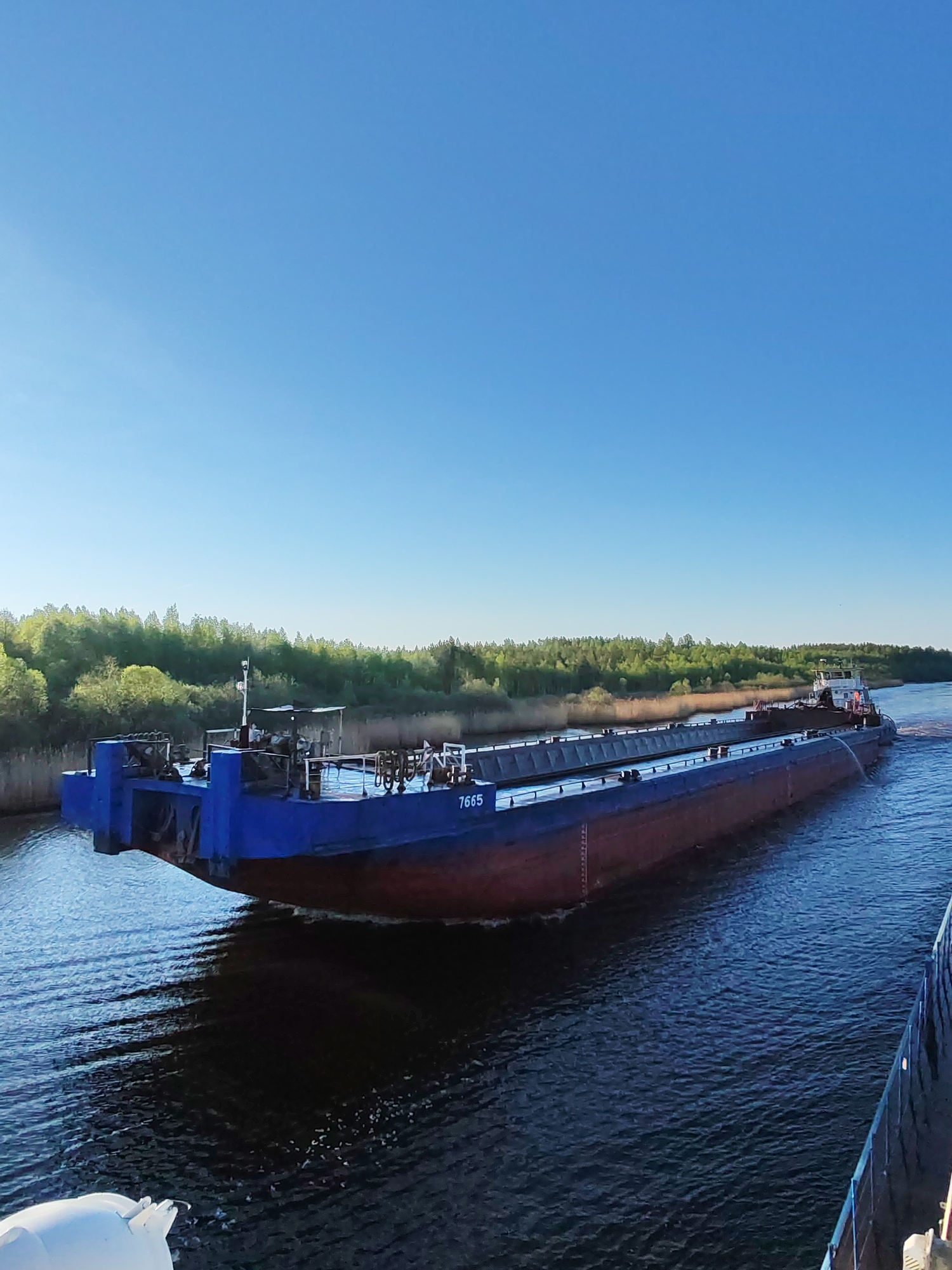 A barge numbered "7665" is underway on a waterway in Ozernoe Ust'e, Russia. The barge has a blue and red hull, a flat top deck, and superstructure sections at both ends. The water is a dark blue, and trees line the shore in the background. The sky is clear and blue. The barge is generating a wake. The edge of another ship is visible in the bottom right corner of the image.