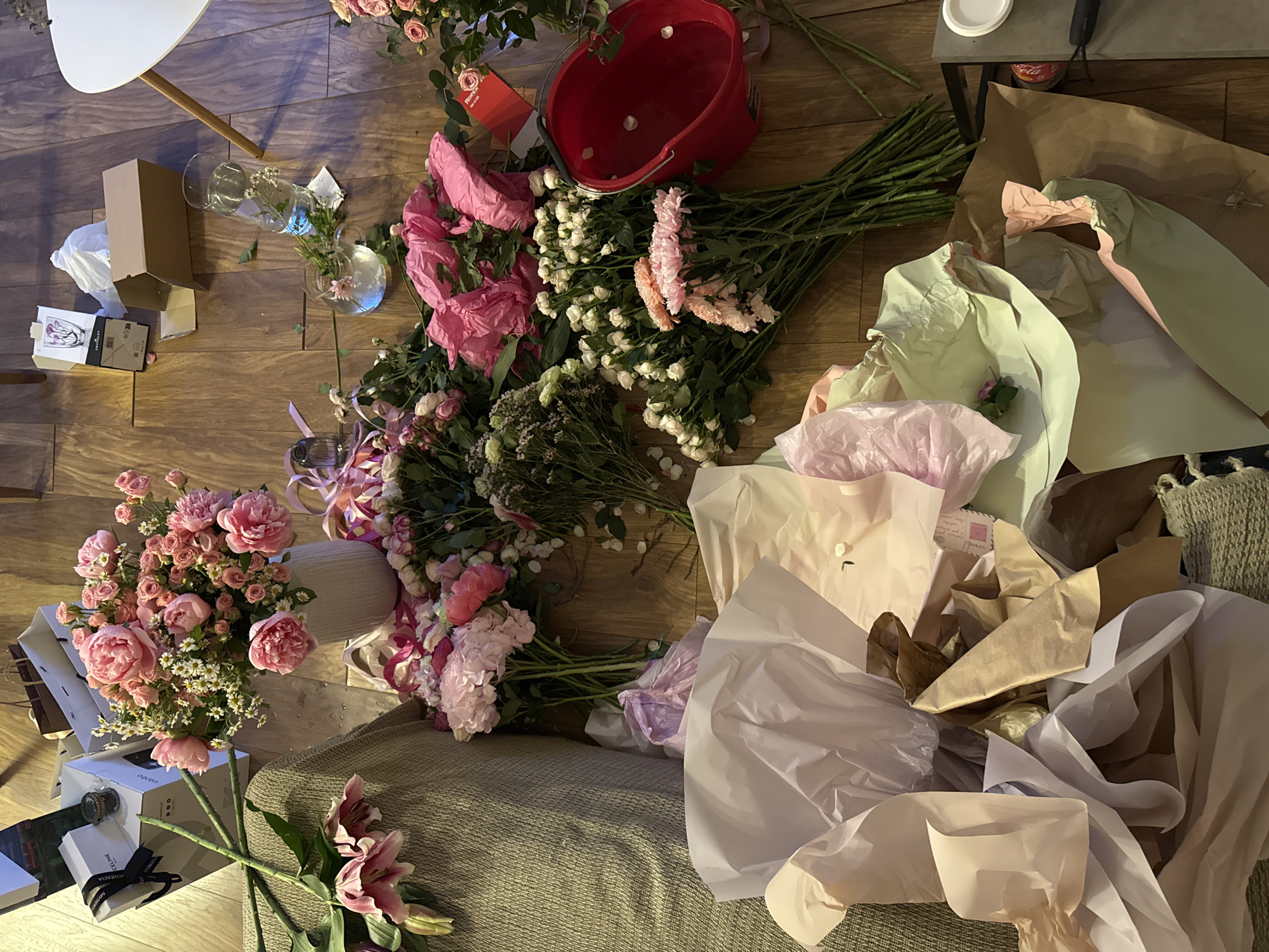 The image shows a collection of flowers and discarded wrapping materials scattered on a floor. Several bouquets of pink, white, and purple flowers are visible, along with a red bucket containing water. Assorted wrapping papers and boxes surround the flowers, suggesting recent unwrapping. A section of a white table and a portion of a brown textured surface, potentially a rug or couch, are also visible. The location appears to be indoors, likely a home.