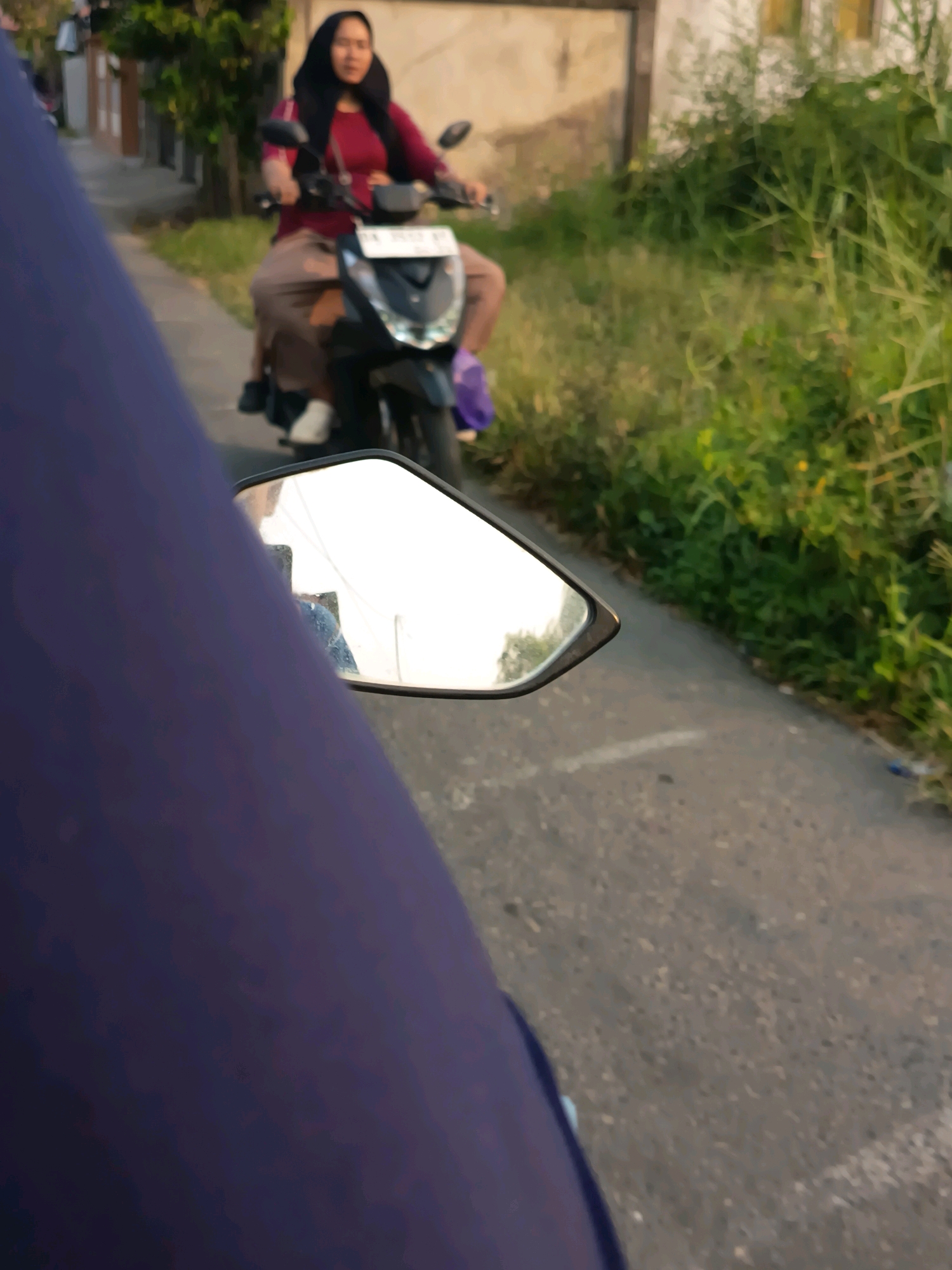 A young woman wearing a black hijab and a maroon shirt rides a black motorcycle along a paved road. She wears brown pants and white shoes. A purple bag is attached to the back of the motorcycle. The motorcycle's side mirror reflects another person on a motorcycle, also wearing a blue shirt. The scene appears to be in a suburban area of Banjarmasin, Indonesia, with a narrow sidewalk and lush green vegetation bordering the road. The lighting suggests it is late afternoon or early evening.