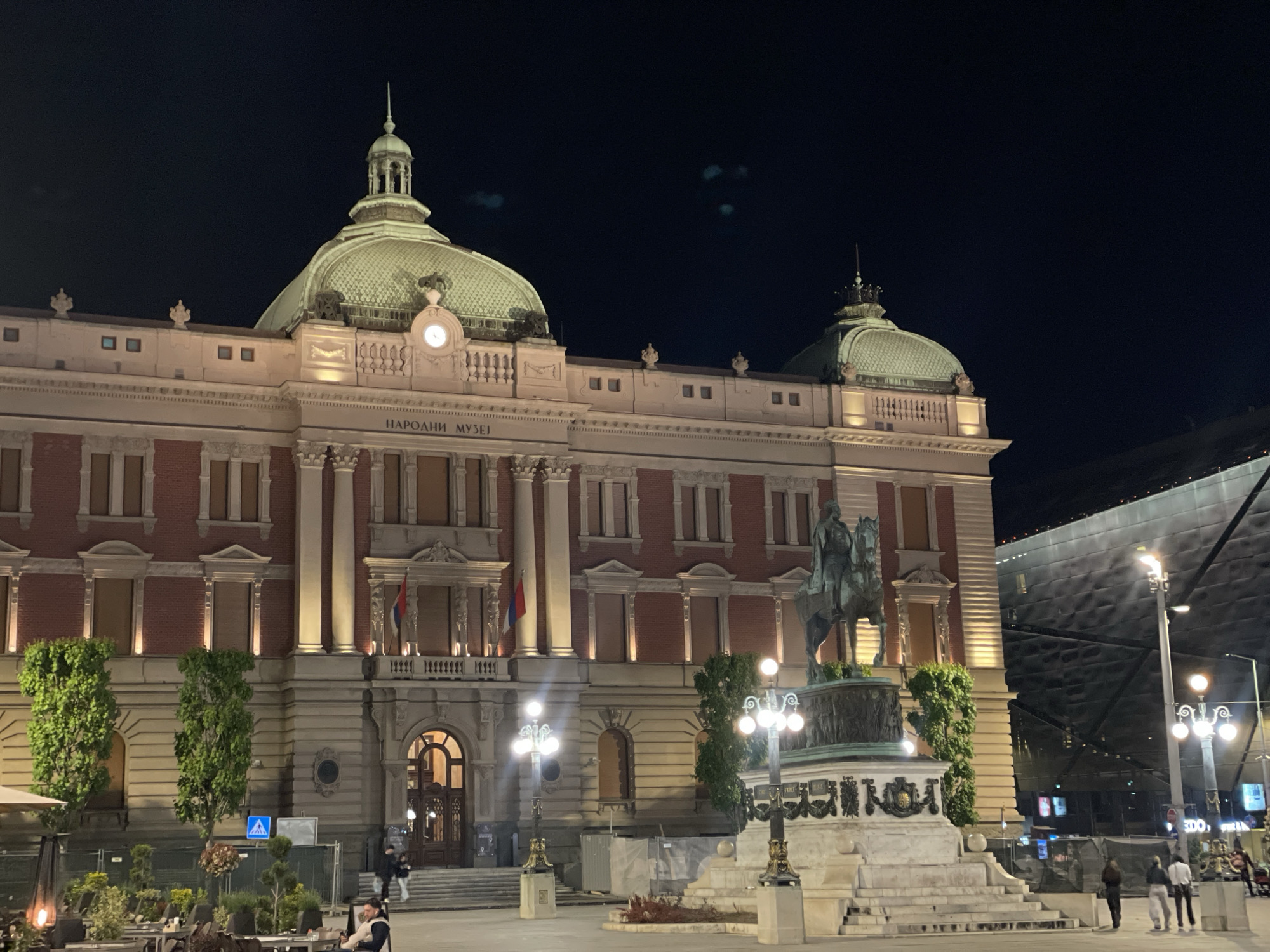 The image shows the National Museum of Serbia in Belgrade at night. The building is illuminated, highlighting its architecture with domes and detailed facades. In front of the museum is the monument to Prince Michael, depicting him on horseback. People are walking around the square. There are some outdoor cafe tables with people sitting at them. The overall scene is vibrant and urban, with a mix of historic architecture and modern city life.