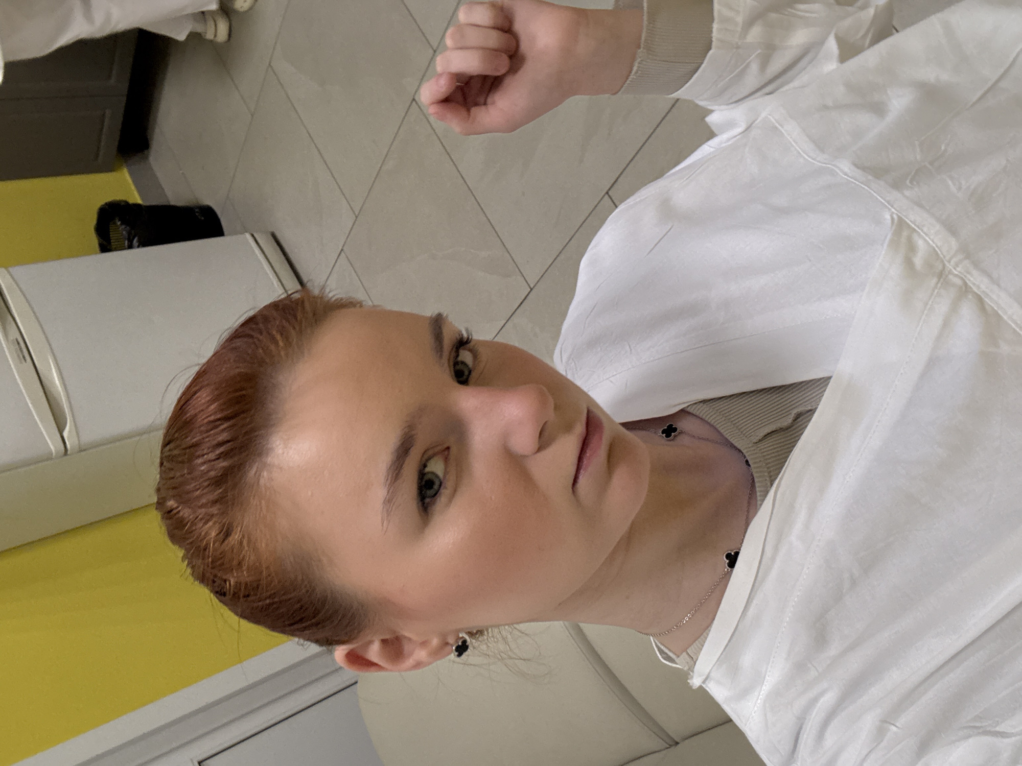 A medium shot depicts a fair-skinned woman with red hair and blue eyes, wearing a white lab coat and a silver necklace with black clover-shaped pendants. She is looking directly at the camera with a neutral expression and a slightly open mouth. Her right hand is visible, held up in a fist. She is also wearing clover-shaped earrings.
The background shows a tiled floor, a white refrigerator with a yellow accent, and a black object on top of it. The location is suggested to be in Unknown, Russia.