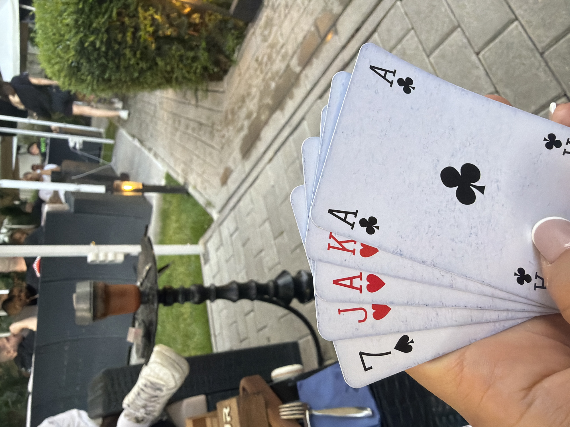 The image captures an outdoor leisure setting, likely a restaurant or lounge patio. In the immediate foreground, a light-skinned hand with manicured nails holds five playing cards fanned out: a 7 of Spades, a Jack of Hearts, an Ace of Hearts, a King of Hearts, and an Ace of Clubs. To the left of the hand, a portion of a brown wooden object, a silver fork, and a blue textile (possibly a napkin or tablecloth) are visible.
In the midground, a black hookah with an amber-colored bowl and a metallic top sits on an ornate black stand. Behind the hookah, a dark grey barrier or partition separates the immediate area from a grass lawn. To the left, part of a white sneaker is visible, suggesting someone is seated nearby.
The background features a paved pathway made of grey rectangular pavers, which curves gently to the right. Several individuals are present, some seated at dark outdoor tables behind the grey barrier, while one person in dark clothing and white sneakers walks away from the camera on the paved path. White vertical poles, possibly supporting an overhead structure, are distributed throughout the background. Artificial lighting includes a warm-glowing lantern on a short pole to the left of the path and another light source embedded within a dense, trimmed green bush on the right side of the path. The overall atmosphere suggests a relaxed, possibly evening, social gathering.
While the request specifies Sofia, Bulgaria, the image itself contains no discernible landmarks, signs, or unique architectural features to definitively confirm this precise geographical location.