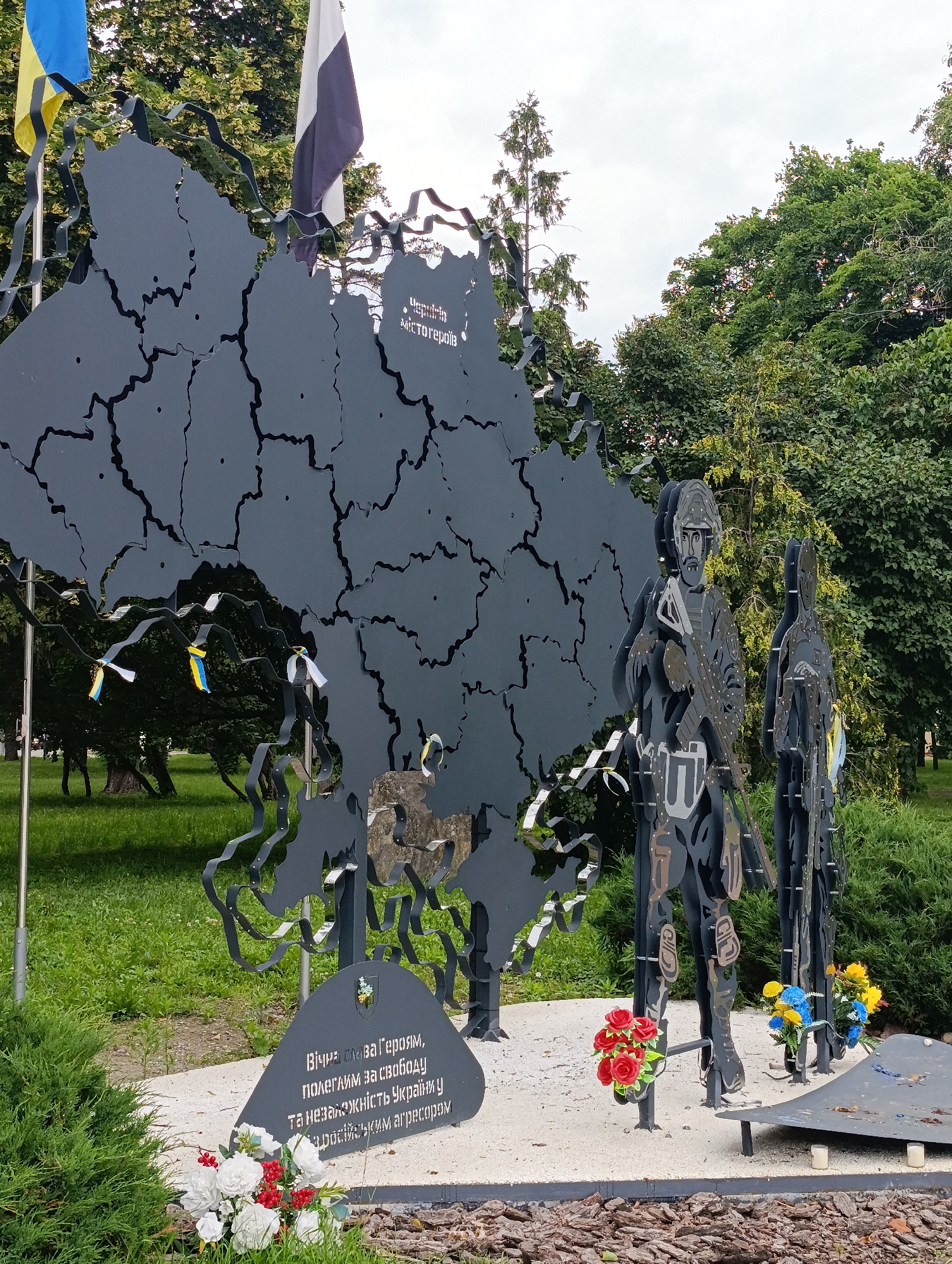 The image depicts a war memorial in Chernihiv, Ukraine, featuring a large, dark metal cutout of the map of Ukraine with scattered circular perforations representing shelling. Two silhouetted figures of soldiers stand in front of the map, one holding a rifle. Ukrainian flags and ribbons are affixed to the metal structures and scattered around the base. In the foreground, a stone plaque with Ukrainian text and a coat of arms is visible, accompanied by a bouquet of white and red artificial flowers. Another bouquet of blue and yellow artificial flowers is placed near the soldiers. The memorial is situated outdoors in a grassy area with trees in the background, under an overcast sky.