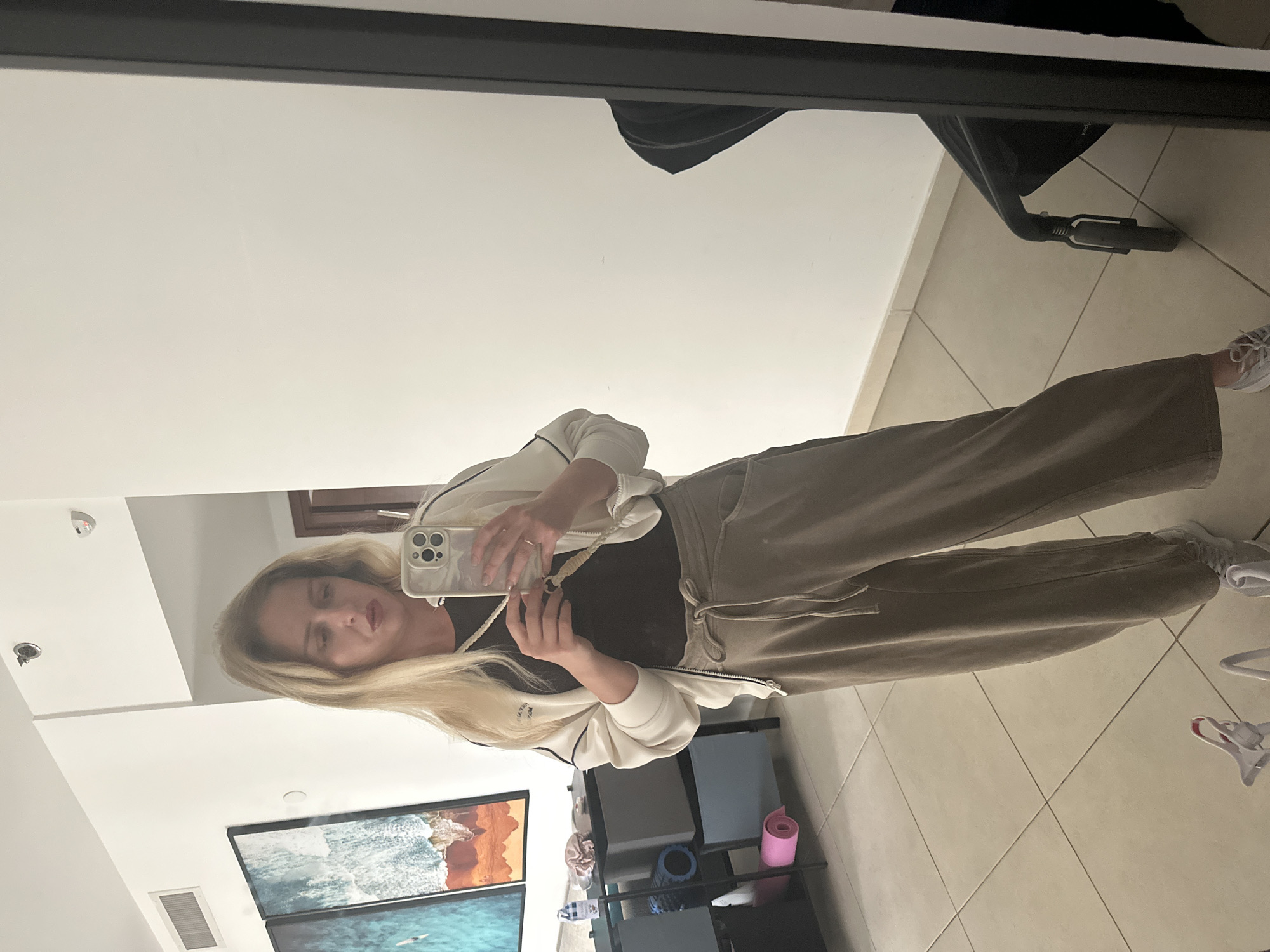 A mirrored reflection shows a person with long blonde hair holding a smartphone to take a picture. The person is wearing a black top, a white jacket, and brown pants. The reflection shows a tiled floor and a wall with a framed picture of a beach scene. A shelf with various items, including a pink mat and blue rollers, is visible. A baby stroller is also partially visible in the reflection. The location is Petah Tikva, Israel.