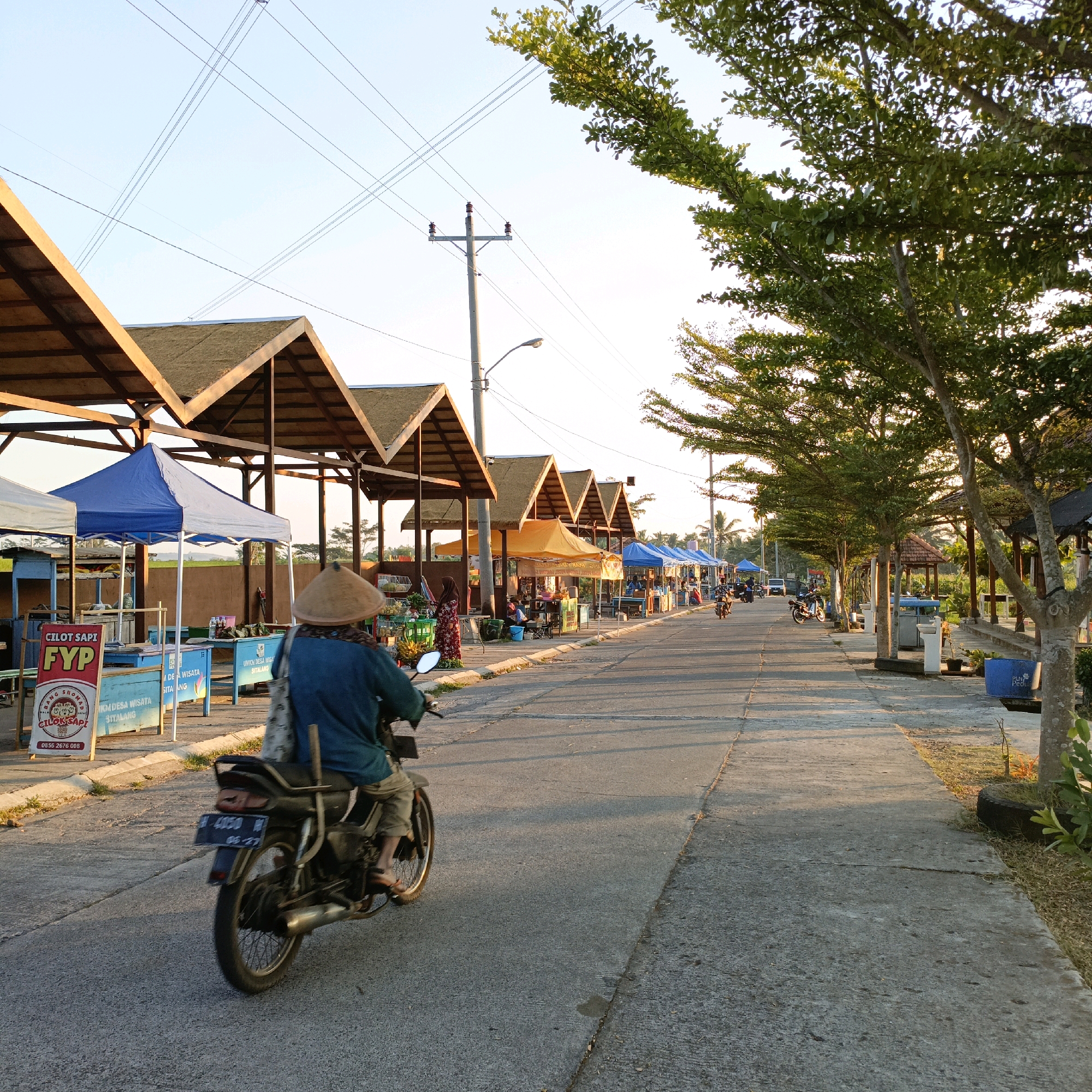 In this image, taken at the Wisata Sitalang tourist area in Salatiga, Indonesia, a person on a motorbike is riding down a paved road. The rider, wearing a conical hat and a blue shirt, is moving away from the camera towards a row of market stalls. These stalls have peaked, thatched roofs supported by wooden beams, and some are covered with blue and orange tarps. Beyond the stalls, there are trees lining the right side of the road, their leaves illuminated by the setting sun. Power lines crisscross the clear sky. The overall scene depicts a local market or roadside commercial area in a tropical environment during the late afternoon.