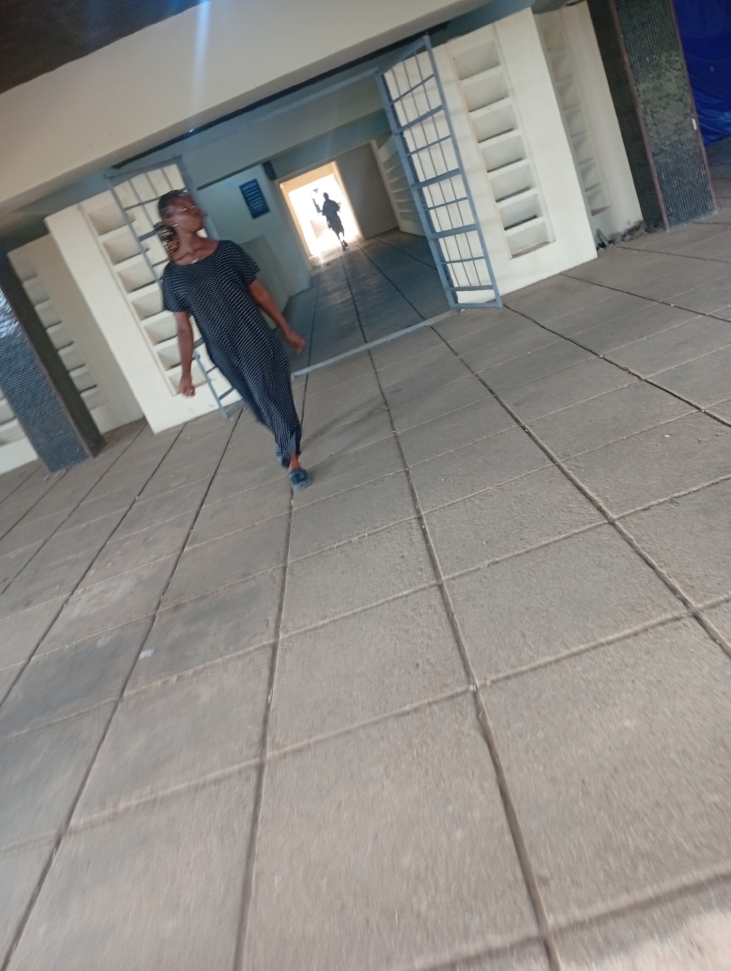 The image shows a woman in a black and white patterned dress standing on a tiled walkway. She is looking to her left. In the background, there is an open doorway with a silhouette of a person walking through it. The environment appears to be an outdoor walkway with a building on the left featuring shelves or cubbies. This scene is likely located in Jos, Nigeria, given the context provided. The walkway is made of large, rectangular tiles. The lighting suggests it is daytime.