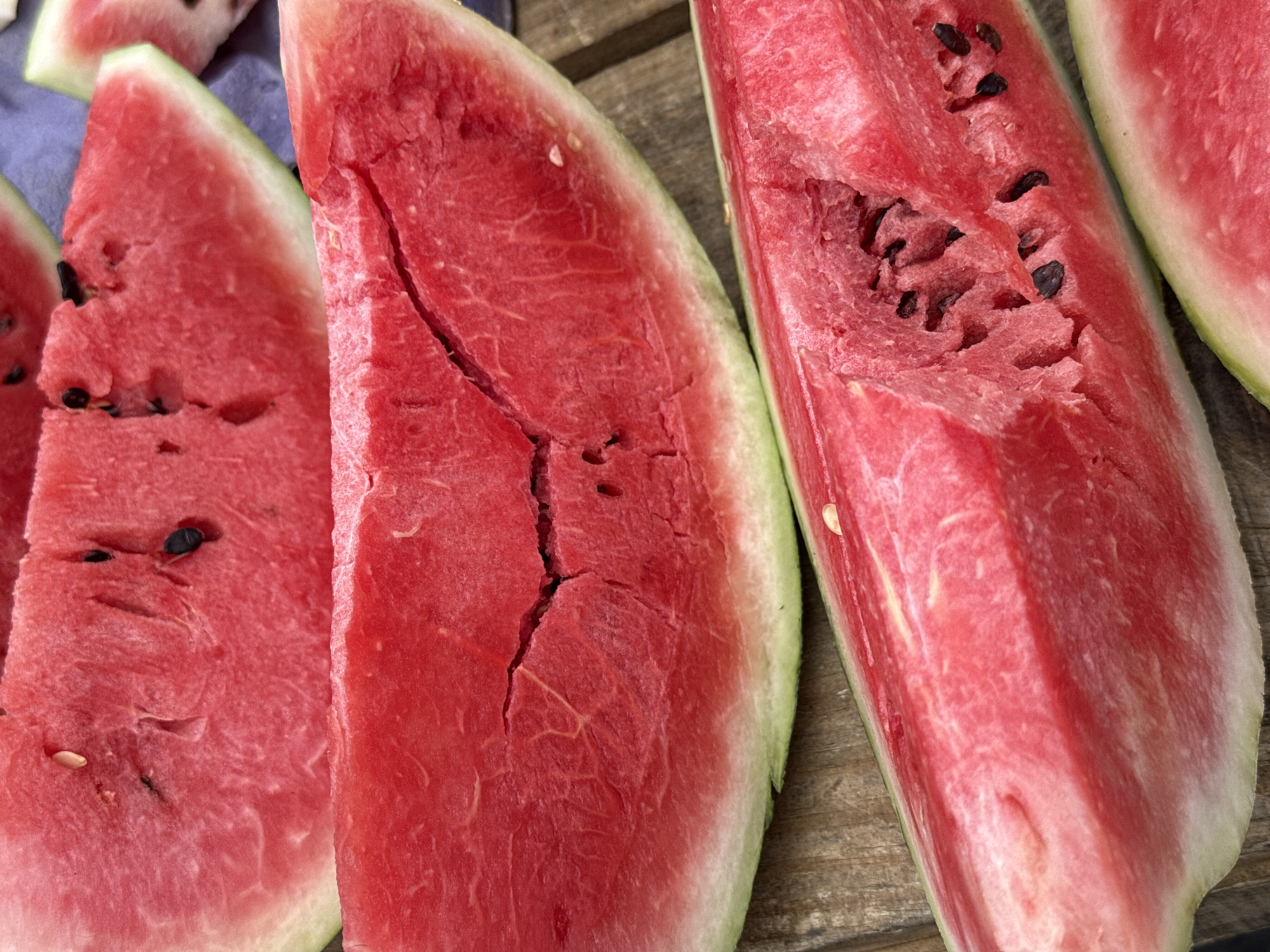 A vertical close-up, high-angle shot shows several slices of watermelon on a wooden surface. The watermelon slices are a vibrant red, with some showing black seeds and a thin green rind. The slices are arranged diagonally, with the top slice being triangular and the bottom two slices being elongated and curved. The lighting is bright, highlighting the texture and juiciness of the fruit. The scene is likely a casual outdoor setting, possibly a backyard or a park, where someone is enjoying a refreshing snack. The environment appears to be clean and well-lit. The image does not contain any people or animals.