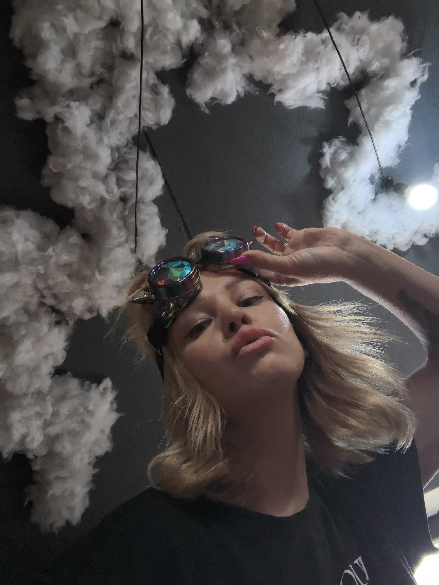 An overhead shot captures a young woman posing indoors in Noviy, Russia, looking up at the camera. She has blonde, wavy hair that frames her face, and her lips are puckered in a "duck face" expression. Distinctive steampunk-style goggles with iridescent, faceted lenses are pushed up onto her forehead. Her right hand is raised near her head, fingers slightly splayed, showcasing bright pink nail polish. She wears a dark, likely black, t-shirt with some unreadable white text on the chest.
The background features a dark gray or black ceiling, heavily adorned with numerous clusters of white, fluffy material, resembling artificial clouds or cotton. A single, bare lightbulb hangs from the ceiling on the right side, casting light and partially obscured by the "clouds." A thin black wire or cord is visible across the ceiling amidst the cloud decor. The scene suggests an indoor location, possibly a themed venue or an artistically decorated space, with artificial lighting creating a unique atmosphere. The subject's pose and accessories contribute to a playful and stylish mood.
