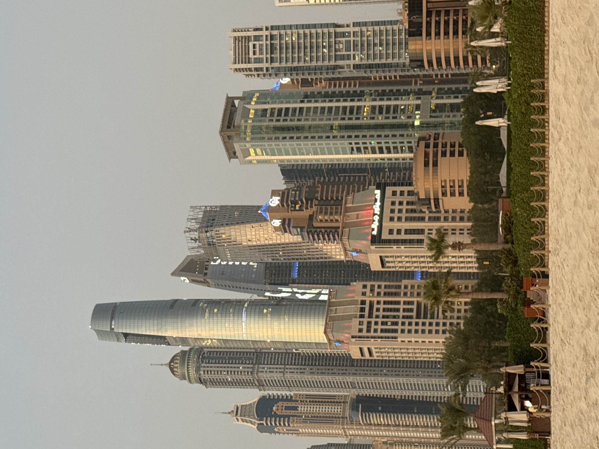 This is a photograph of the Dubai Marina skyline, taken from a sandy beach. The image captures a dense collection of skyscrapers with varying architectural styles, some clad in glass and others in stone. The tallest building, a curved, glass skyscraper, dominates the center of the frame, its facade reflecting the soft, overcast sky. To its left, a distinctive tower with a domed top is visible. Several other high-rise buildings, some with intricate facades and illuminated windows, are interspersed throughout the cityscape.
In the foreground, a beach area is depicted. A low rope barrier demarcates the edge of the sand. Behind the barrier, there is a lush green hedge, punctuated by palm trees. To the left of the frame, a small, open-air structure with a brown, conical roof is present, featuring several white umbrellas and seating arrangements, suggesting a beachside cafe or lounge area. A few individuals are visible within or near this structure. White beach umbrellas are also scattered along the green area. The overall lighting suggests it is either early morning or late afternoon, with a muted, diffused light. The sky is a uniform light gray, devoid of any distinct clouds or the sun.