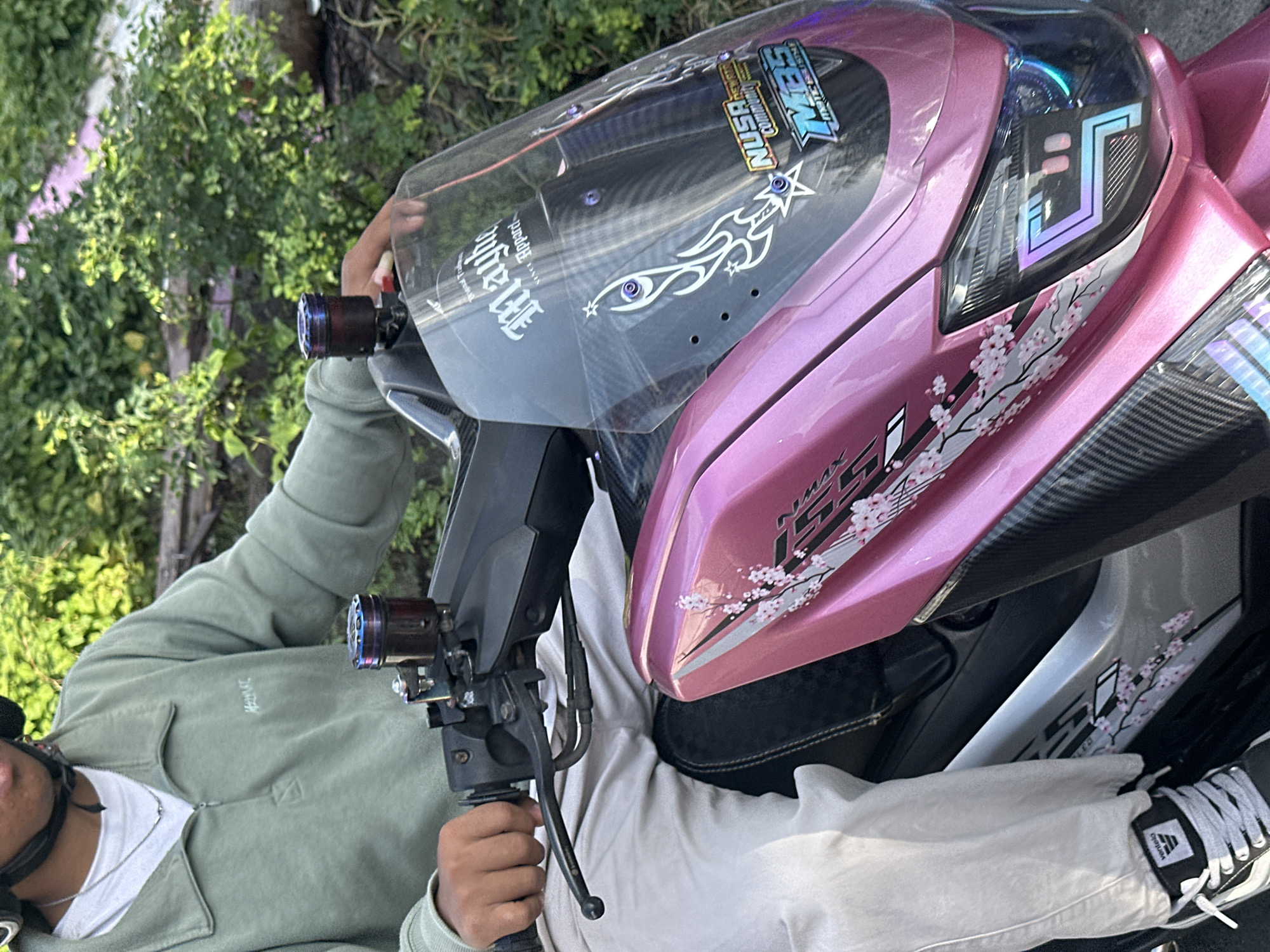 In North Kuta, Indonesia, a person wearing a light green quarter-zip sweatshirt, white pants, and black and white sneakers is seated on a pink and silver scooter. The scooter has a clear windshield with decals, including a flame-like design, a star, and stickers that read "NUSA", "COMMUNITY", and "M35 MATIC STYLE". The scooter also features carbon fiber accents and pink cherry blossom decals on its body. The person is holding the handlebars, and a black helmet is visible on their head. The background consists of lush green foliage.