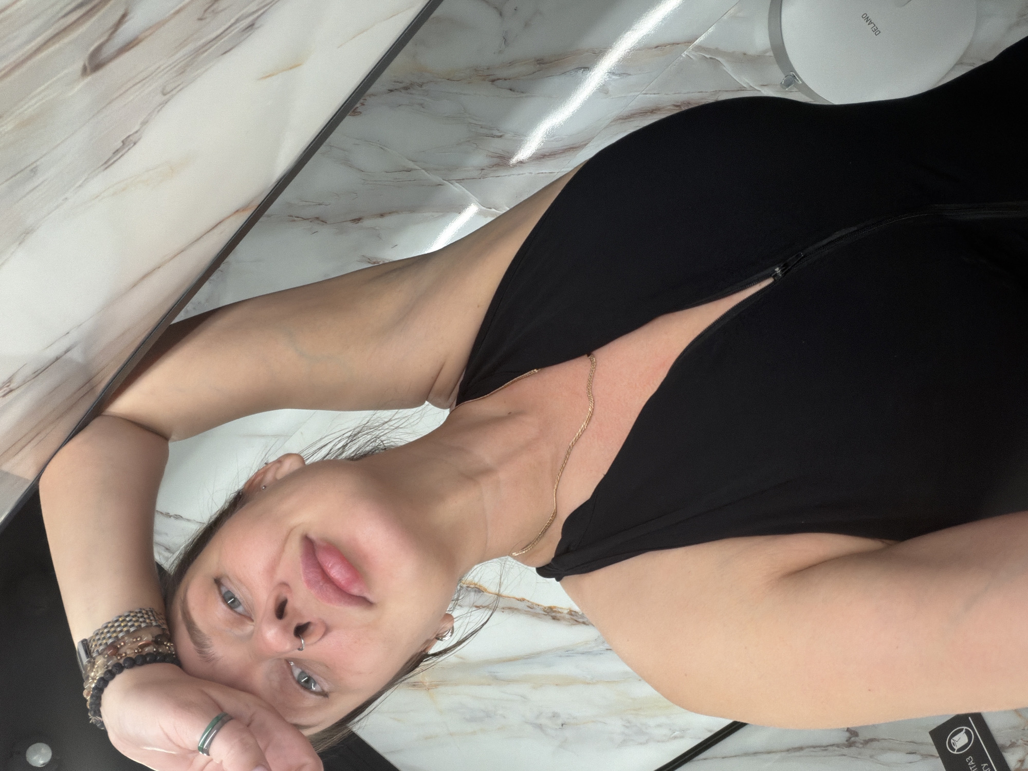 A woman lies on a marble surface wearing a black dress and a gold necklace. She has a nose ring and is wearing multiple bracelets on her left wrist. A Delano product is visible in the upper right corner. The location cannot be determined with the given information; there is no indication it is Moscow, Russia.