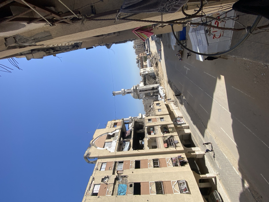 This image depicts a street scene in Khan Yunis, Palestinian Territory, under a clear blue sky. The perspective is elevated, looking down a paved street flanked by damaged buildings.
On the left, a multi-story, light-colored building exhibits significant structural damage, with upper floors missing walls, exposing interiors, and showing damaged balconies. Some windows are covered with blue tarps, and clothes hang from several balconies. A car is parked on the sidewalk at the base of this building. Two individuals, appearing to be young males, are visible on the sidewalk in the bottom-left, one bending down while the other stands nearby.
The street, marked with white lines and a directional arrow, recedes into the background. In the mid-ground, a severely damaged mosque with a white minaret stands prominently, its main structure partially collapsed. Rubble and debris are scattered around the mosque and extending into the background. Other partially damaged buildings are visible further down the street.
On the immediate right, a partial view of a building's balcony or window frame is visible, with a striped towel and dark fabric hanging. Exposed rebar protrudes from the top edge of this structure. Further down the street on the right, a white tent-like structure with Arabic text is present. Several small figures (people) are sparsely distributed along the street and sidewalks in the mid-ground. Strong shadows are cast on the street and buildings, indicating direct sunlight.