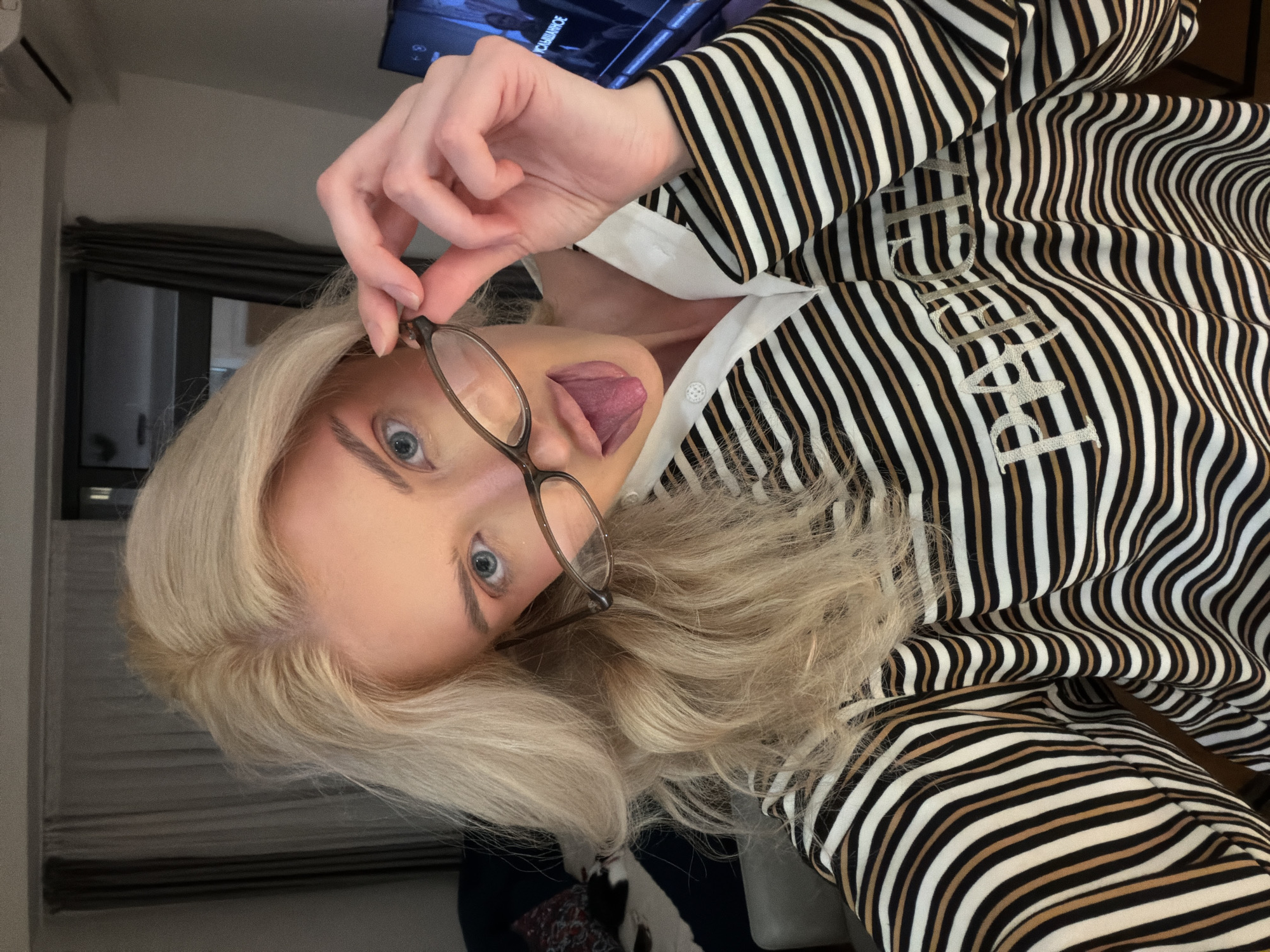 A blonde-haired woman with glasses is sticking her tongue out in an indoor setting. She is wearing a striped shirt with writing on it, seemingly in a private residence. The background includes a window, curtains, and a television screen. There is no visible evidence to suggest this is in Belgrade, Serbia.