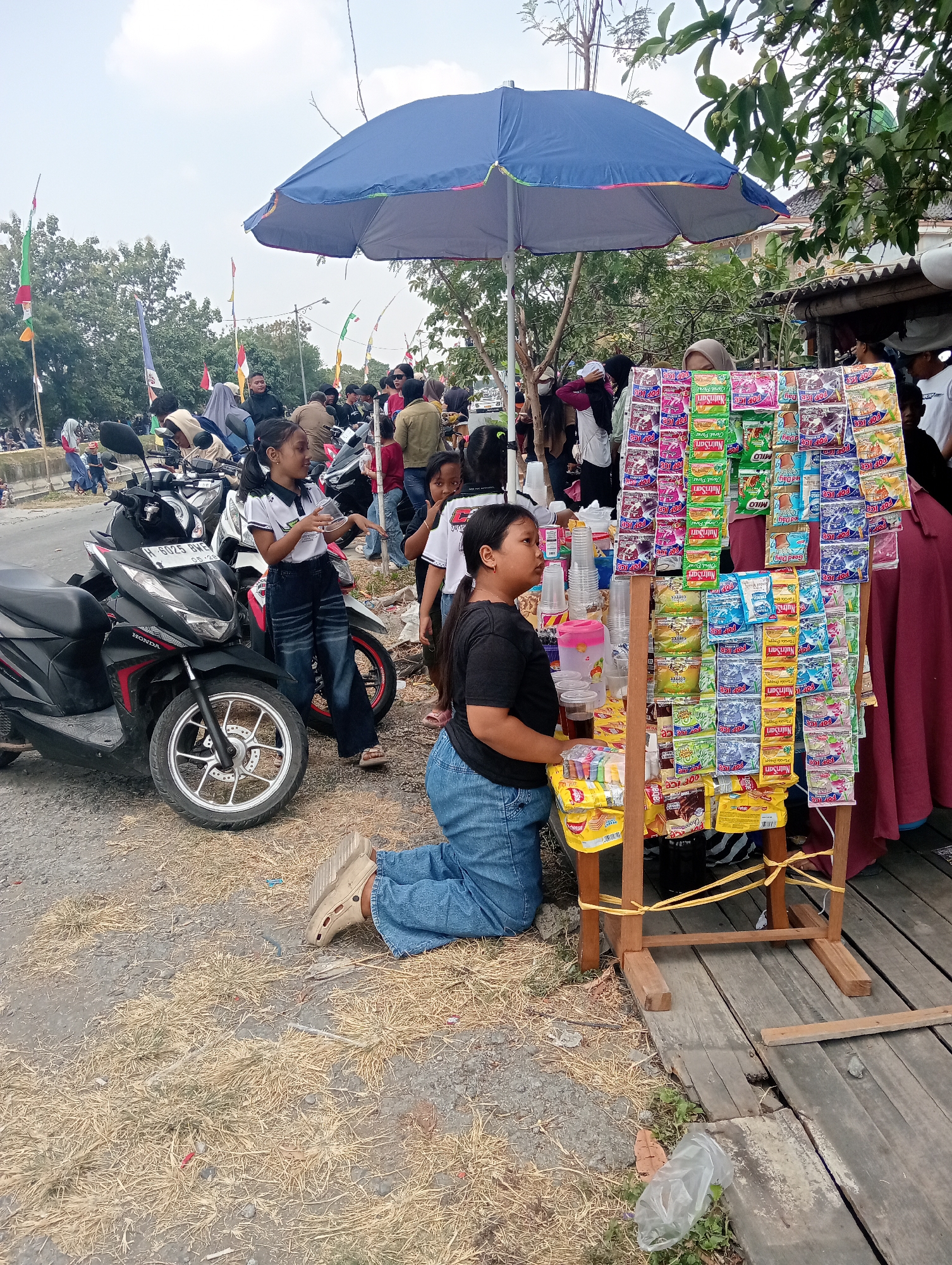 A group of people, including children, are gathered around a street vendor's stall in Doreng, Indonesia. The vendor is selling a variety of packaged snacks and drinks, displayed on a wooden stand under a large blue umbrella. Two young girls are prominently featured: one is kneeling in front of the stall, holding a drink, and another is standing beside motorcycles, looking towards the stall. Other individuals, some on motorcycles, are visible in the background, suggesting a busy street or market environment. The ground is covered with dry grass and gravel, with a wooden walkway in the foreground. Flags are visible in the background, adding to the festive atmosphere.