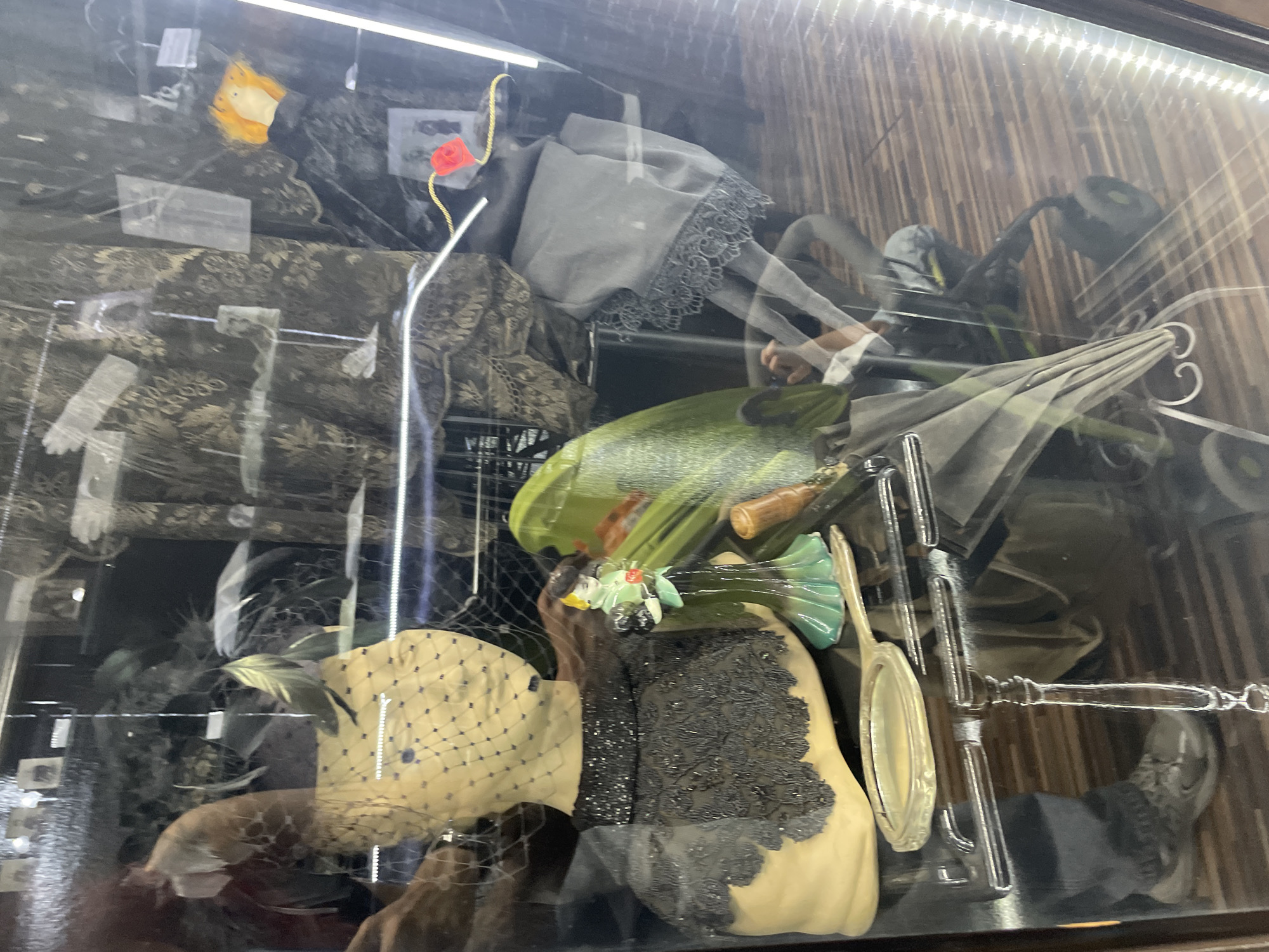 This image captures a display case in a shop, likely in Voskhod, Russia, showcasing vintage fashion accessories. The primary subject is a mannequin bust adorned with a veiled headdress and a heavily beaded black collar and bodice. To the right of the bust, a small figurine of a woman in a green dress holding flowers stands on a delicate stand. A polished silver hand mirror rests in front of the bust. In the background, various textiles with intricate patterns are visible, along with what appear to be framed photographs or cards. A bright green, somewhat abstract object is positioned behind the mannequin. The floor beneath the display is wooden and paneled. The overall impression is of a carefully curated collection of historical items.