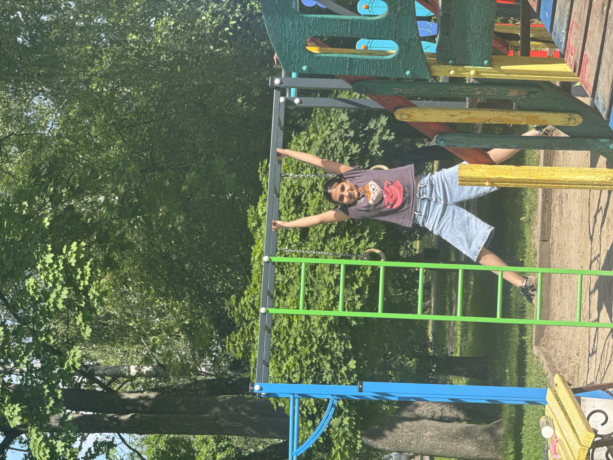 A young person is hanging from a metal bar on a colorful playground structure, with their arms outstretched. They are wearing a dark t-shirt with a cartoon character on it and light blue denim shorts. Their feet are dangling and one leg is bent at the knee. The playground has elements of green, yellow, red, and blue. Behind the playground, there are lush green trees and grass, suggesting an outdoor, possibly park-like setting. The lighting indicates it is daytime, with sunlight casting shadows. The person's posture and expression, though partially obscured, suggest playfulness and enjoyment. The scene appears to be a bright, sunny day, likely in a summer season, given the greenery. The location is identified as Poltava, Ukraine. No visible text is present in the image.