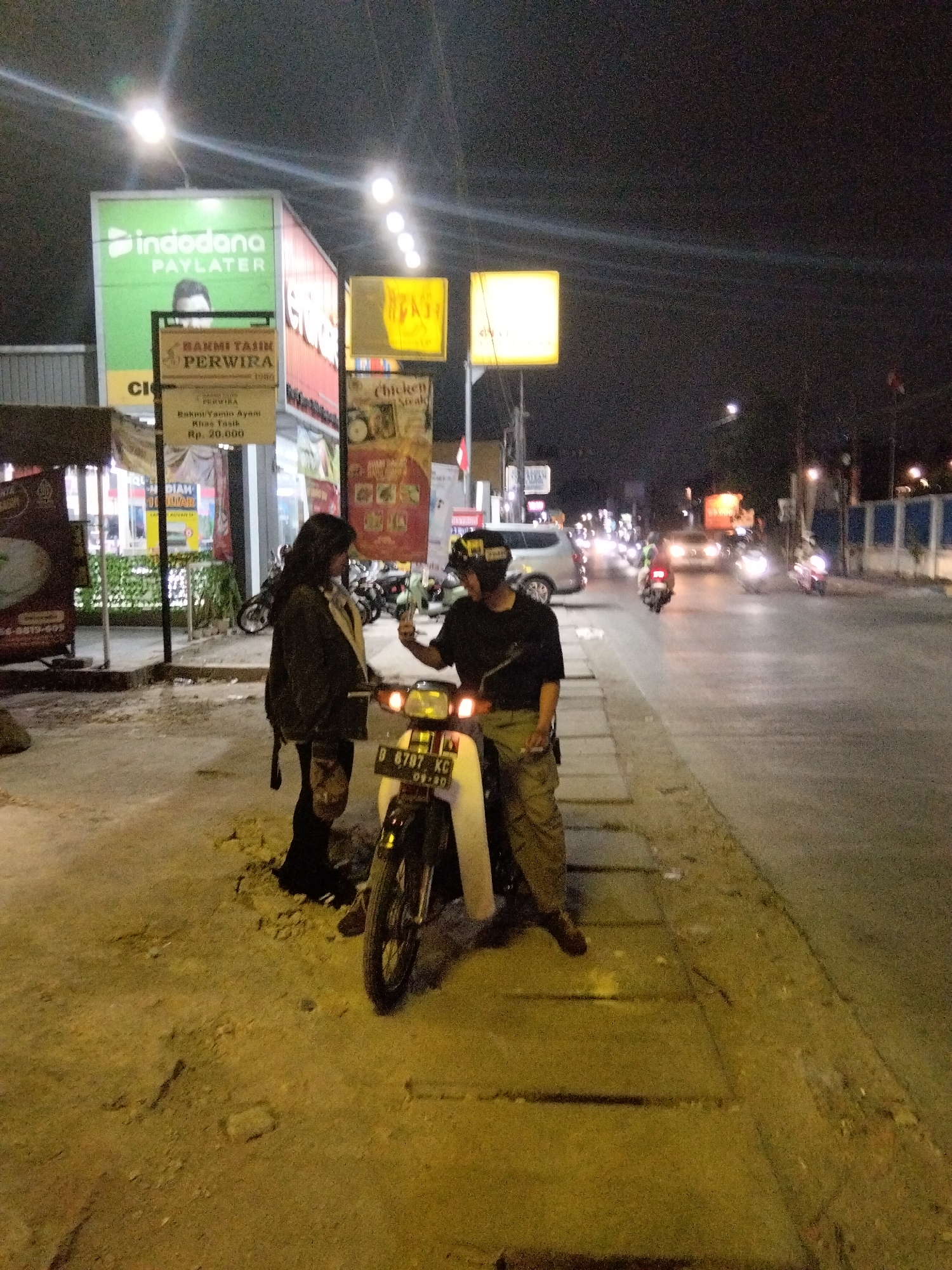 The image captures a nighttime scene on a street in South Tangerang, Indonesia. Two individuals are standing next to a motorcycle parked on the side of a road with a sidewalk. The person on the left, wearing a dark jacket and dark pants, is facing away from the camera, while the person on the right, dressed in a dark t-shirt and khaki pants, is facing the camera, holding a phone. A white motorcycle with license plate B 6787 KG is positioned between them. The background features a street with passing vehicles and illuminated signs. One prominent sign displays "indodana PAYLATER" in green and white. Another sign, featuring red and white, reads "BAKMI TASIK PERWIRA". Further signs for "Chicken Steak" and other businesses are also visible. The street appears to be paved, with a dirt and debris-covered area next to the sidewalk. Streetlights and vehicle headlights illuminate the scene.
