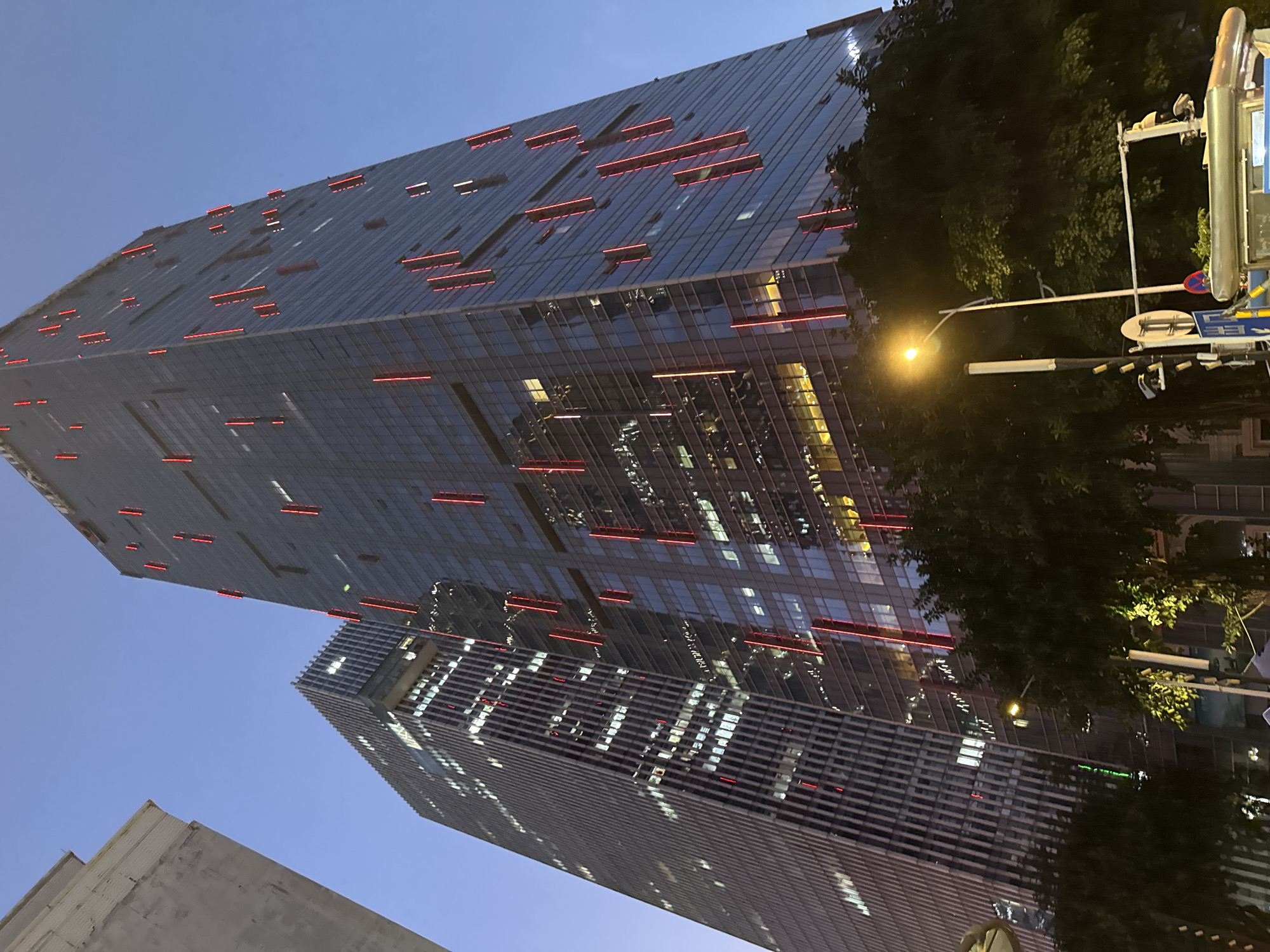 The image shows a nighttime view of modern skyscrapers in Tianhe District, China. Two tall buildings dominate the scene, constructed with glass exteriors. Both buildings feature evenly spaced horizontal red light bars. Trees partially obscure the right side of the frame. A street lamp is visible on the right, providing additional illumination. In the lower left, part of another building is seen. The sky is a clear, dark blue.