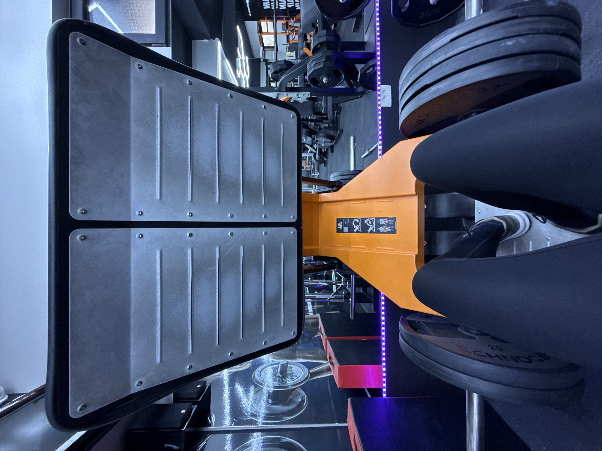 The image captures an indoor scene at a gym in Budva, Montenegro, with a focus on a leg press machine. The machine features two large, rectangular footplates with a gray metallic finish and bolted detailing. The weight loading mechanism is visible, showcasing several black weight plates stacked on a vertical bar. A person wearing black leggings and white and gray sneakers is positioned in the machine, presumably mid-exercise. The gym's interior includes a mirrored wall reflecting other equipment, accent lighting (likely LED strips) with a purplish hue, and patterned flooring. Other gym equipment and structural elements are visible in the background.