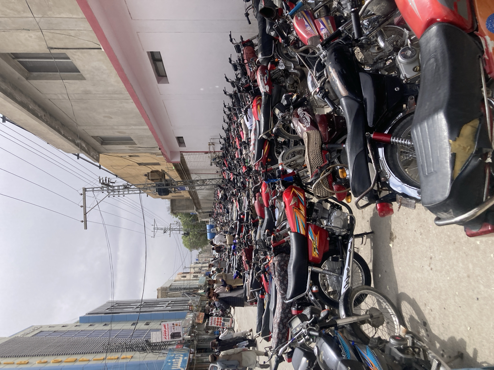 The image depicts a dense concentration of motorcycles parked on a concrete surface in an urban environment. A group of men are standing among the motorcycles. The background reveals buildings, power lines, and a cloudy sky. The location is Quetta City Tehsil, Pakistan. The objects are motorcycles and buildings. The subject is Quetta.