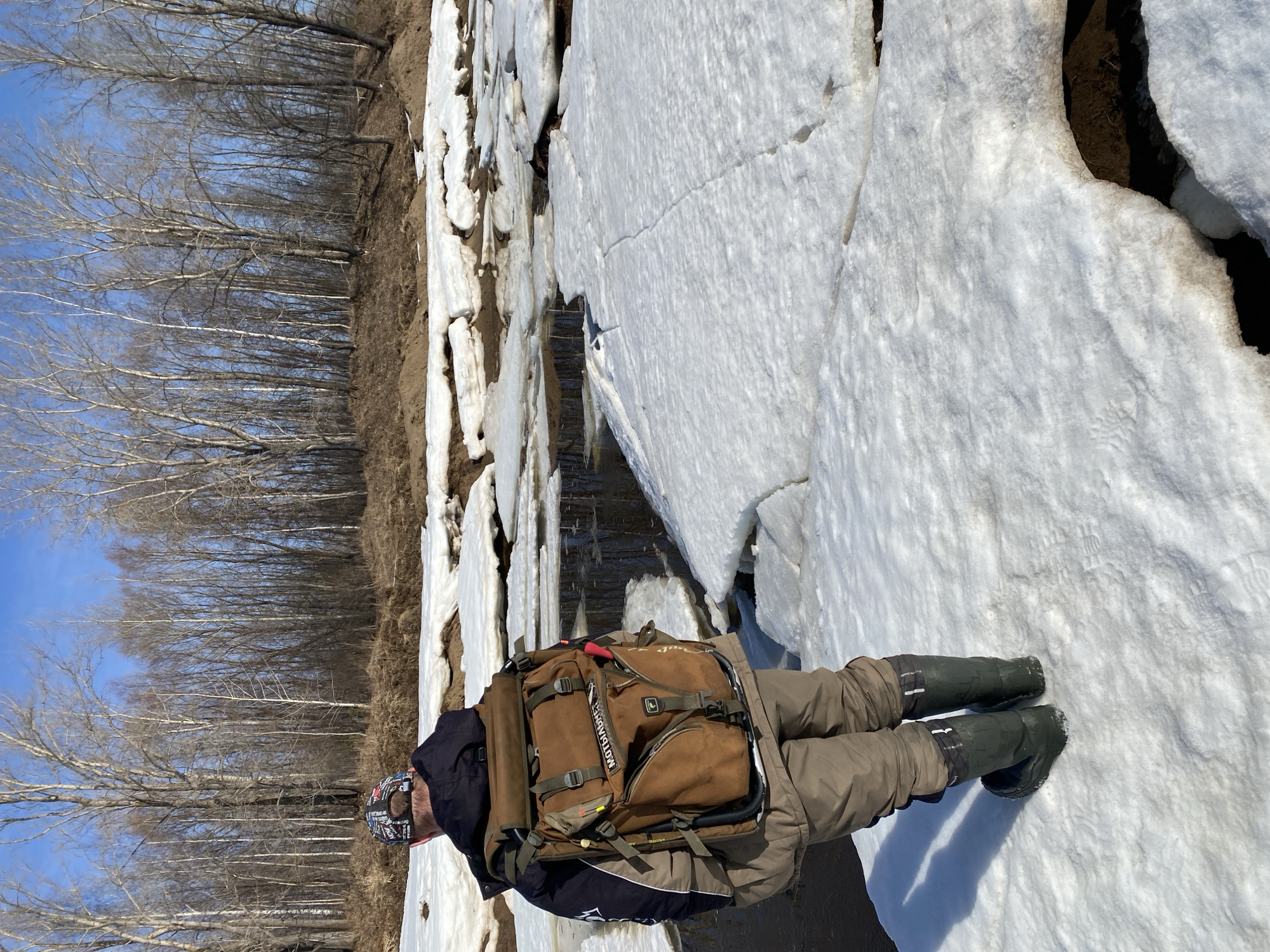 An outdoor scene shows a person wearing a backpack and green boots standing on a sheet of cracked ice and snow. The backdrop includes a forest of bare trees against a blue sky. Water is visible between the cracks in the ice. The person is wearing a dark-colored jacket, and tan pants. The backpack is brown and appears to be a hiking pack. The location is Ol'khovskoye, Russia.