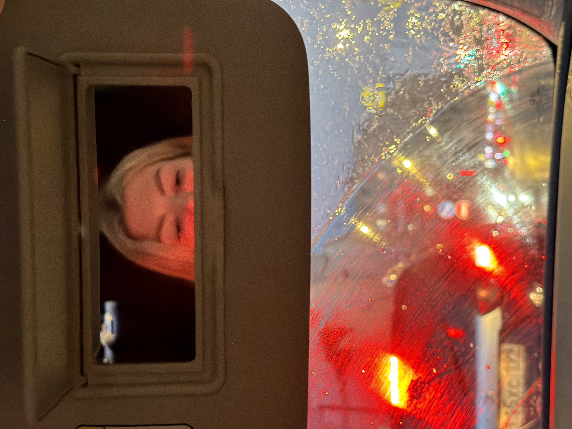The image shows a close-up view inside a vehicle, likely a car, during nighttime with rain. The upper portion of the image features a vanity mirror from the sun visor, reflecting a portion of a woman's face, specifically her eyes and forehead. The reflection is dimly lit, with the woman's blonde hair visible.
The lower portion of the image looks through the car's windshield, which is speckled with raindrops. This view reveals a busy city street scene at night, characterized by numerous blurred lights from other vehicles and traffic signals. The dominant colors are red from brake lights and yellow/white from headlights and streetlights, creating a bokeh effect due to the rain. There is a license plate visible on the car in front, partially obscured by the rain and lights, with characters that appear to be Cyrillic. The overall impression is that of being in traffic in a city at night during a rain shower, with the driver checking their appearance in the mirror. The presence of Cyrillic script on the license plate suggests the location is likely Russia, and potentially Moscow given the urban setting and traffic density.