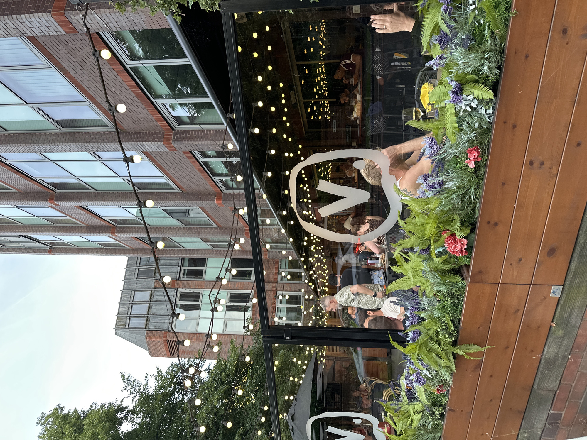 An outdoor dining area of a restaurant is pictured, enclosed by glass walls and decorated with string lights. In the foreground, a wooden planter box is filled with ferns and purple flowers. Through the glass, patrons are seated at tables, some eating and drinking. A large white "V" logo is visible on the glass. In the background, a brick building with large windows rises above. The sky is overcast.