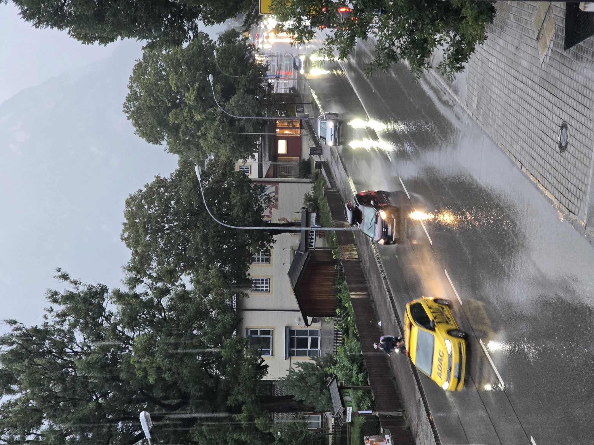 The image shows a street scene in Garmisch-Partenkirchen, Germany, on a rainy day. The road is wet and reflects the lights of the vehicles. Several cars are visible, including a yellow ADAC taxi. A person is riding a motorcycle in the foreground. Trees line the street, and a building with a light-colored facade and a wooden shed is visible on the left. In the background, a mountain is obscured by fog. The overall impression is one of a wet, overcast day in a German town.
