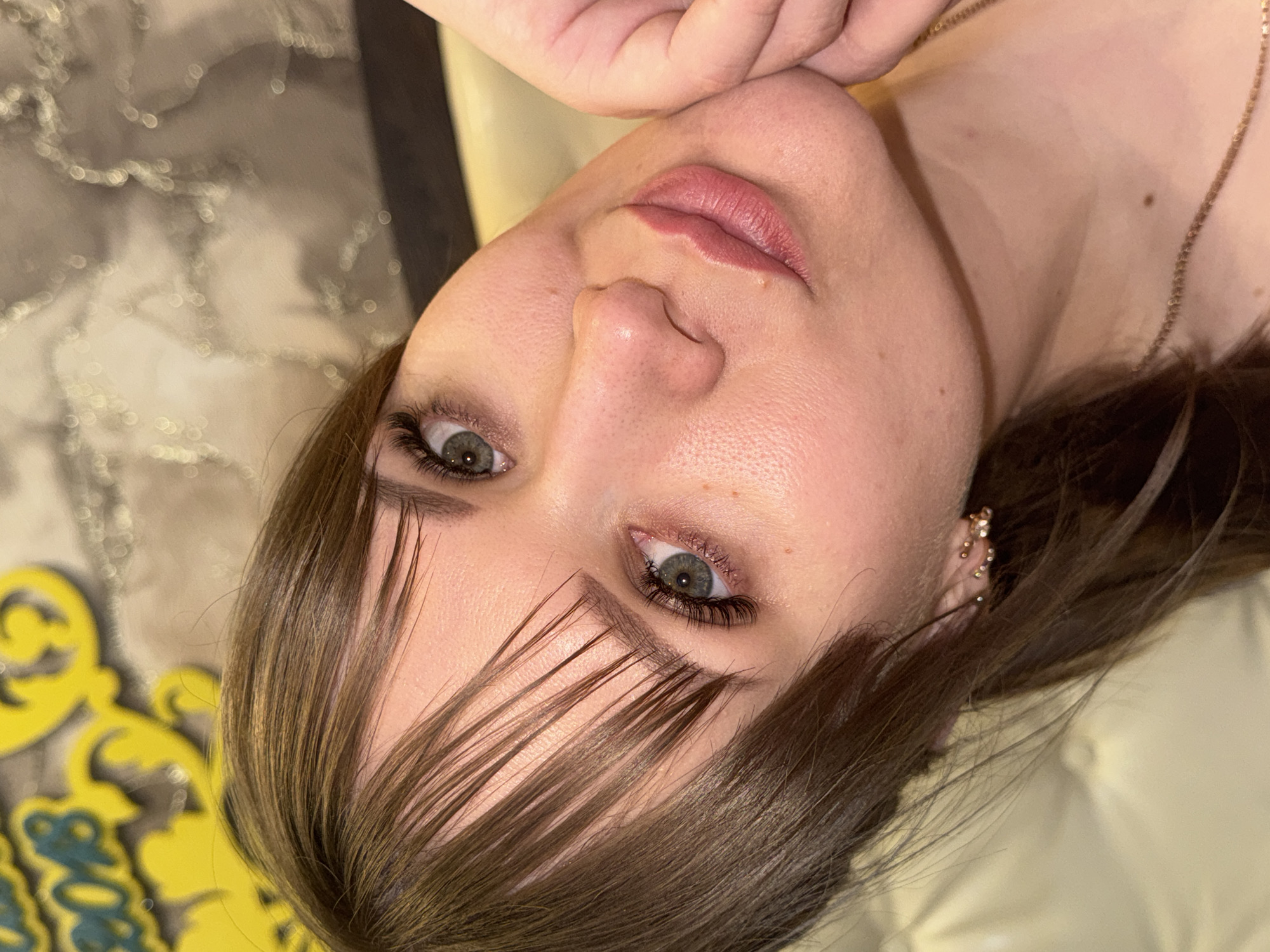 Close-up of a fair-skinned woman with green eyes, light brown hair with bangs partially covering her face, pink lips, and a gold necklace and earrings. She appears to be posing for the camera. The background includes a blurred, yellow-colored object and a textured surface. The location is not discernible as Chaikovsky, Russia based on the provided image.