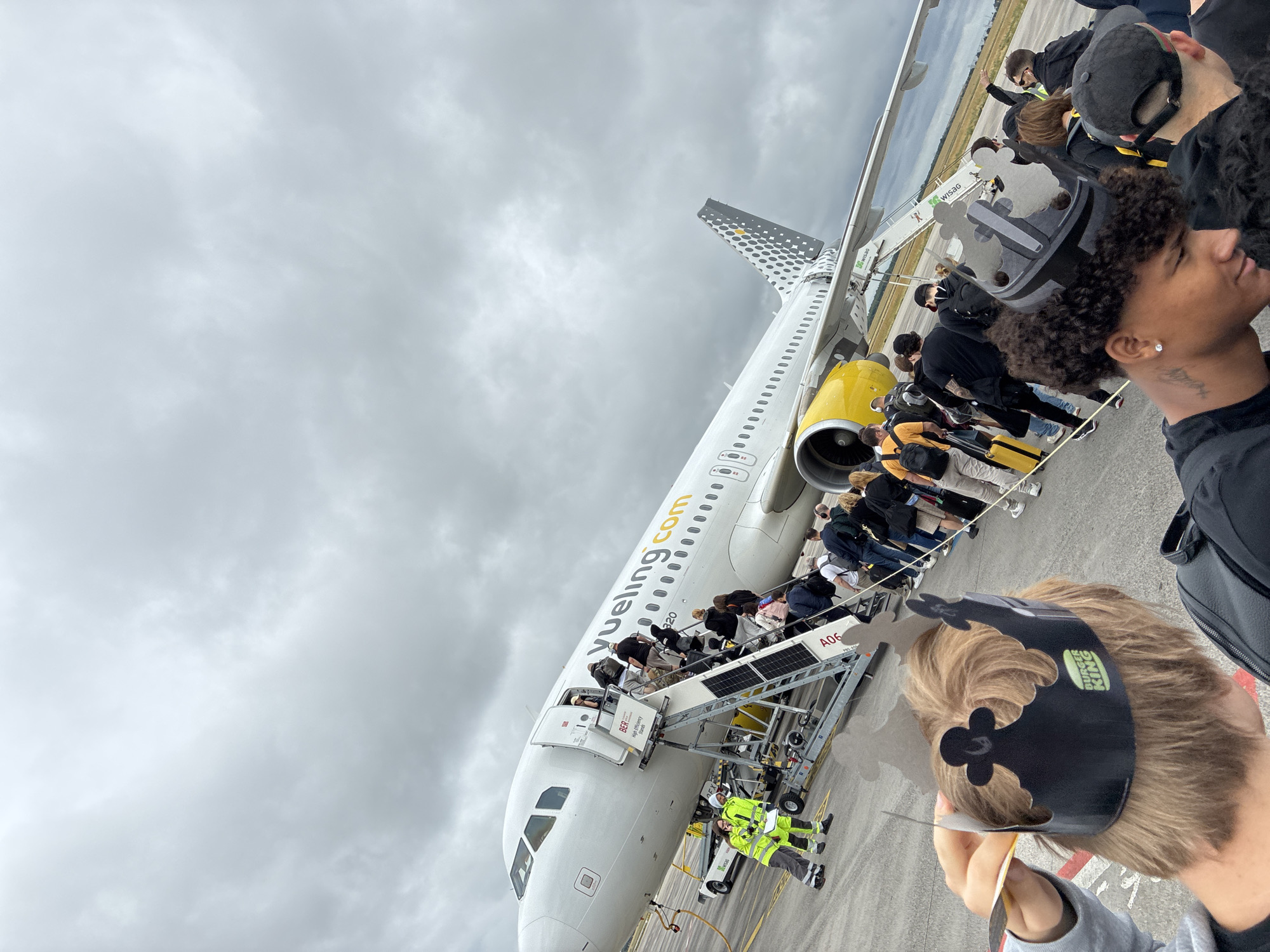 In Schönefeld, Germany, under a cloudy sky, passengers are boarding a Vueling airplane via a mobile staircase. The airplane, with its distinctive yellow and grey livery, is parked on the tarmac. Two ground crew members in high-visibility vests are standing near the staircase. A line of passengers, some carrying luggage, is ascending the stairs. The foreground shows the backs of two individuals, one wearing a Burger King crown, suggesting a casual or playful atmosphere despite the travel context. The overall scene depicts the boarding process at an airport.
