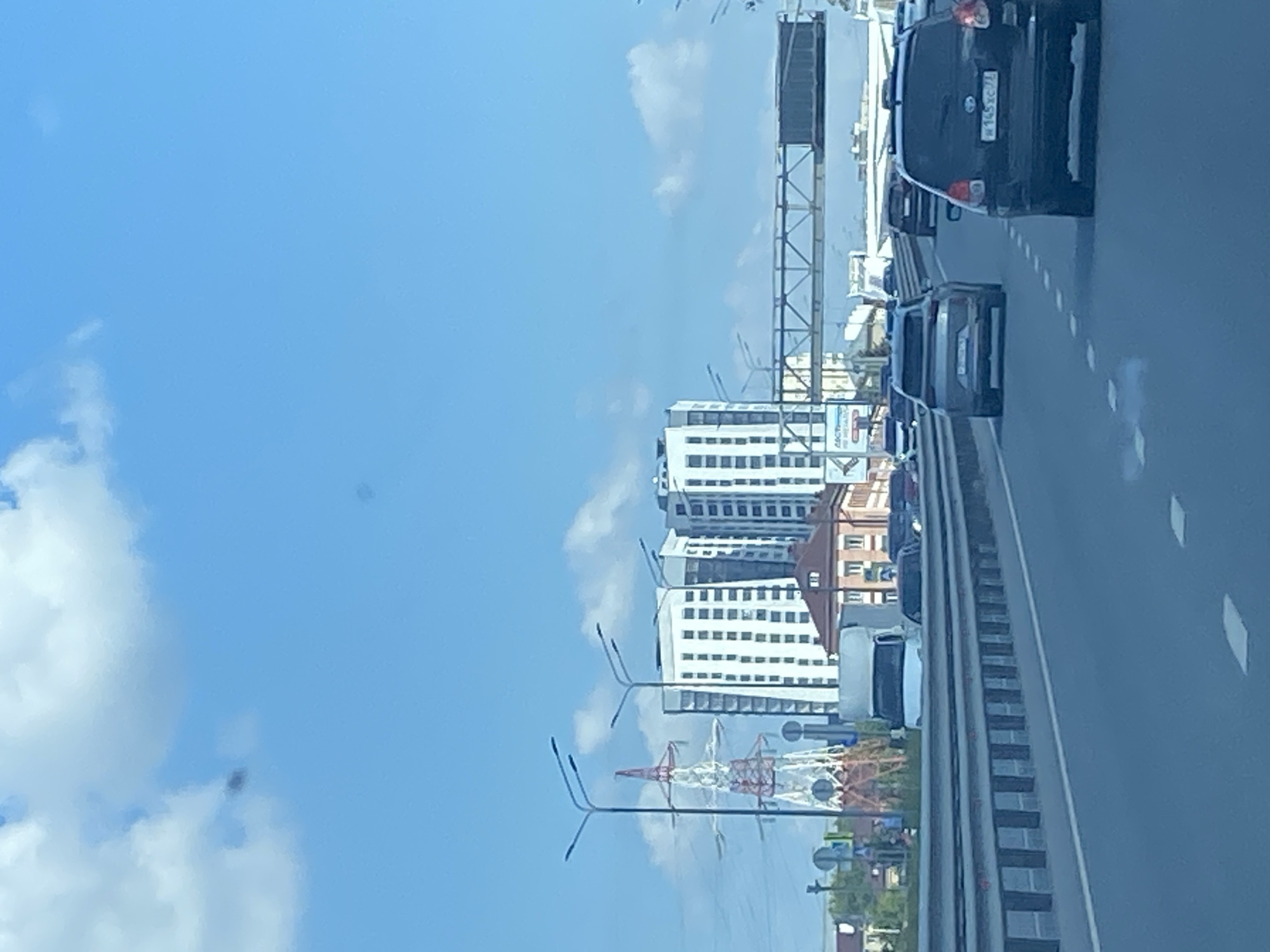 The image depicts a view from a highway in Kazan, Russia. The foreground is dominated by a multi-lane asphalt road with clear white lane markings. Several cars are visible driving on the highway, including a black SUV in the right lane with a Russian license plate reading "W 145 XC 71". To the left of this SUV is a silver SUV. In the distance, a white truck is parked on the shoulder. Beyond the highway, a row of modern, white apartment buildings can be seen, along with a red-roofed building and a prominent red and white lattice pylon, likely a power line tower. The sky is bright blue with scattered white clouds. The overall scene suggests a typical urban environment on a clear day.