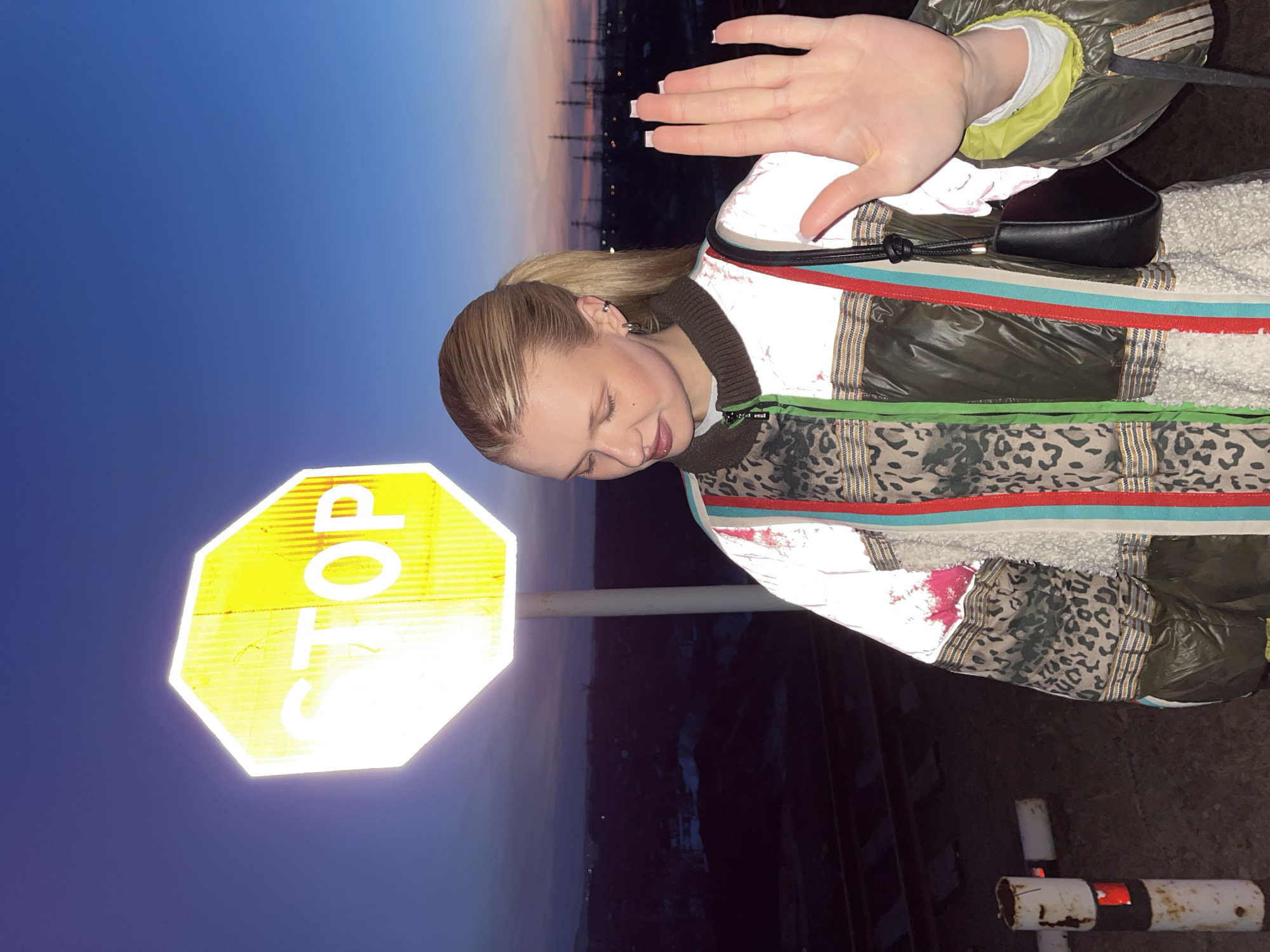 A woman stands next to a stop sign, with a cityscape in the background. She is wearing a patterned jacket and holding her hand up. The image was taken in Chaikovsky, Russia.