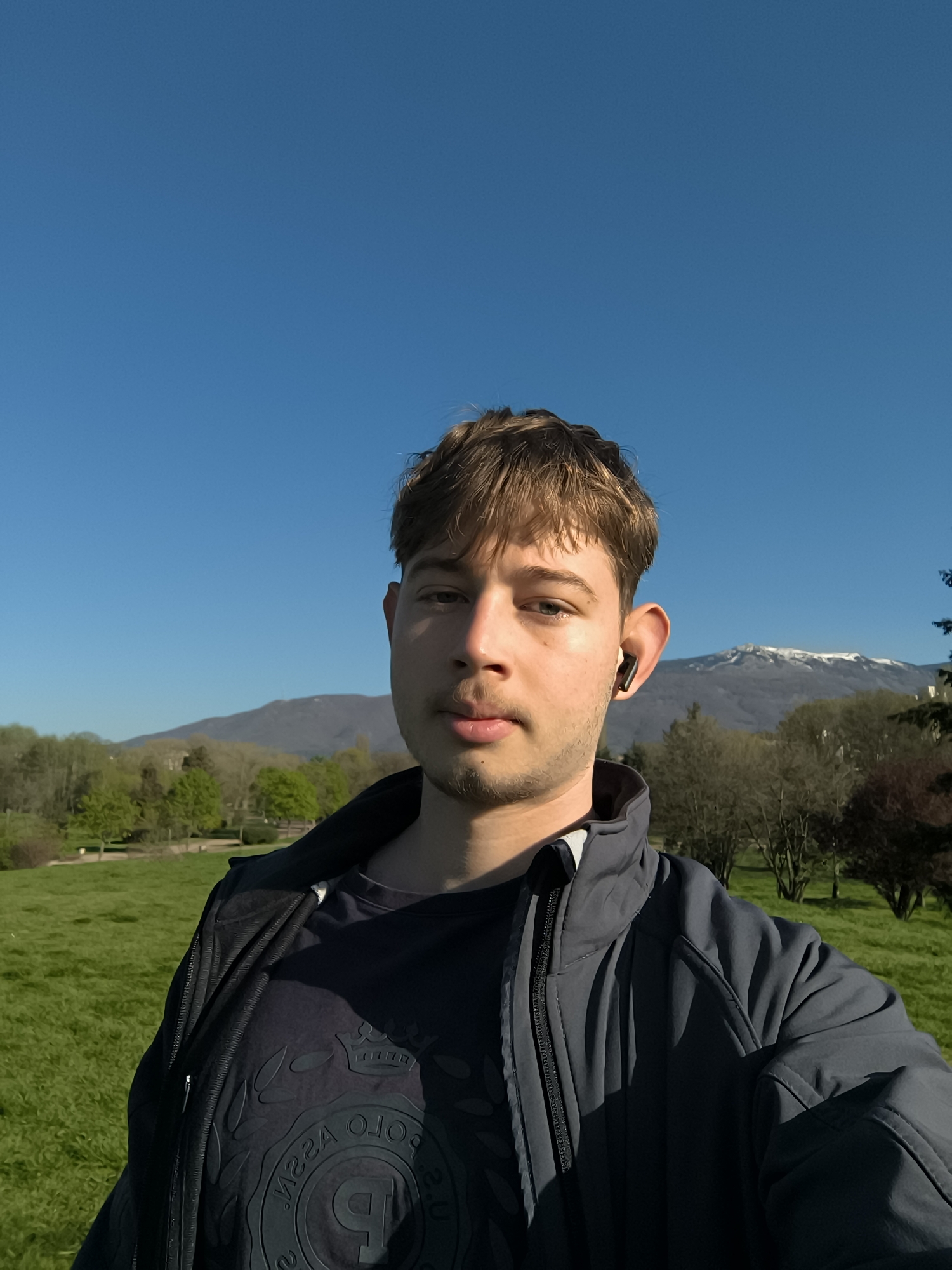 The image is a selfie of a young man with short brown hair, wearing a dark blue T-shirt under a dark grey jacket. He has an earbud in his right ear. The background shows a green park with trees and a mountain range in the distance, under a clear blue sky. The mountain peak has snow on it. Based on the geography, this is likely a park in Sofia, Bulgaria, with the Vitosha mountain range in the background.