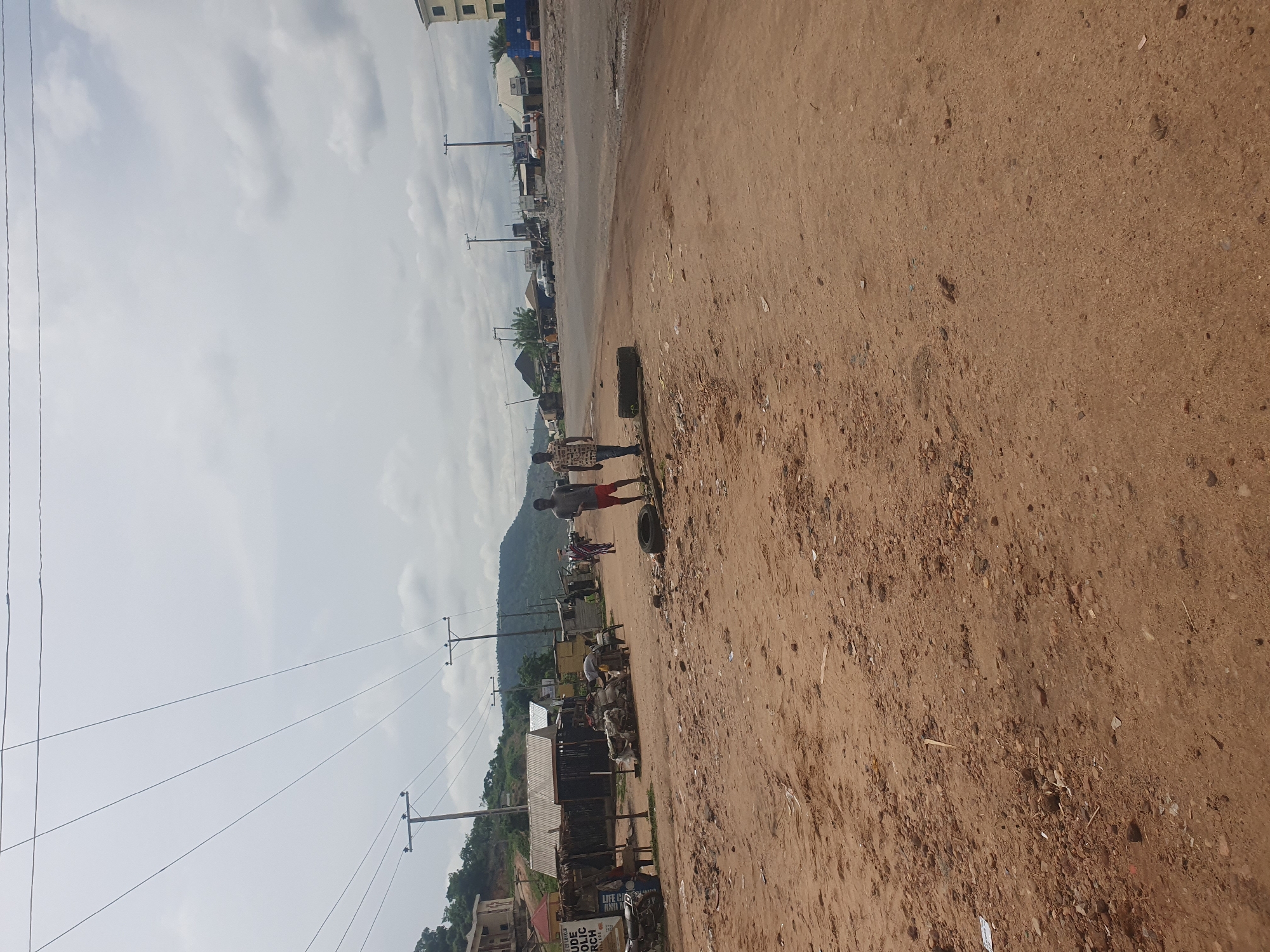The image depicts a dusty street scene in Lokoja, Nigeria. The road is unpaved, primarily composed of reddish-brown soil. Two individuals are visible, carrying what appears to be a flat object between them. One is shirtless, wearing red shorts, and the other has on a patterned shirt. Several discarded tires and debris lie along the roadside. Power lines stretch across the sky, supported by wooden poles. Distant structures and green vegetation are visible against the backdrop of a mountain. A sign for a church is partly visible. The sky is cloudy.
