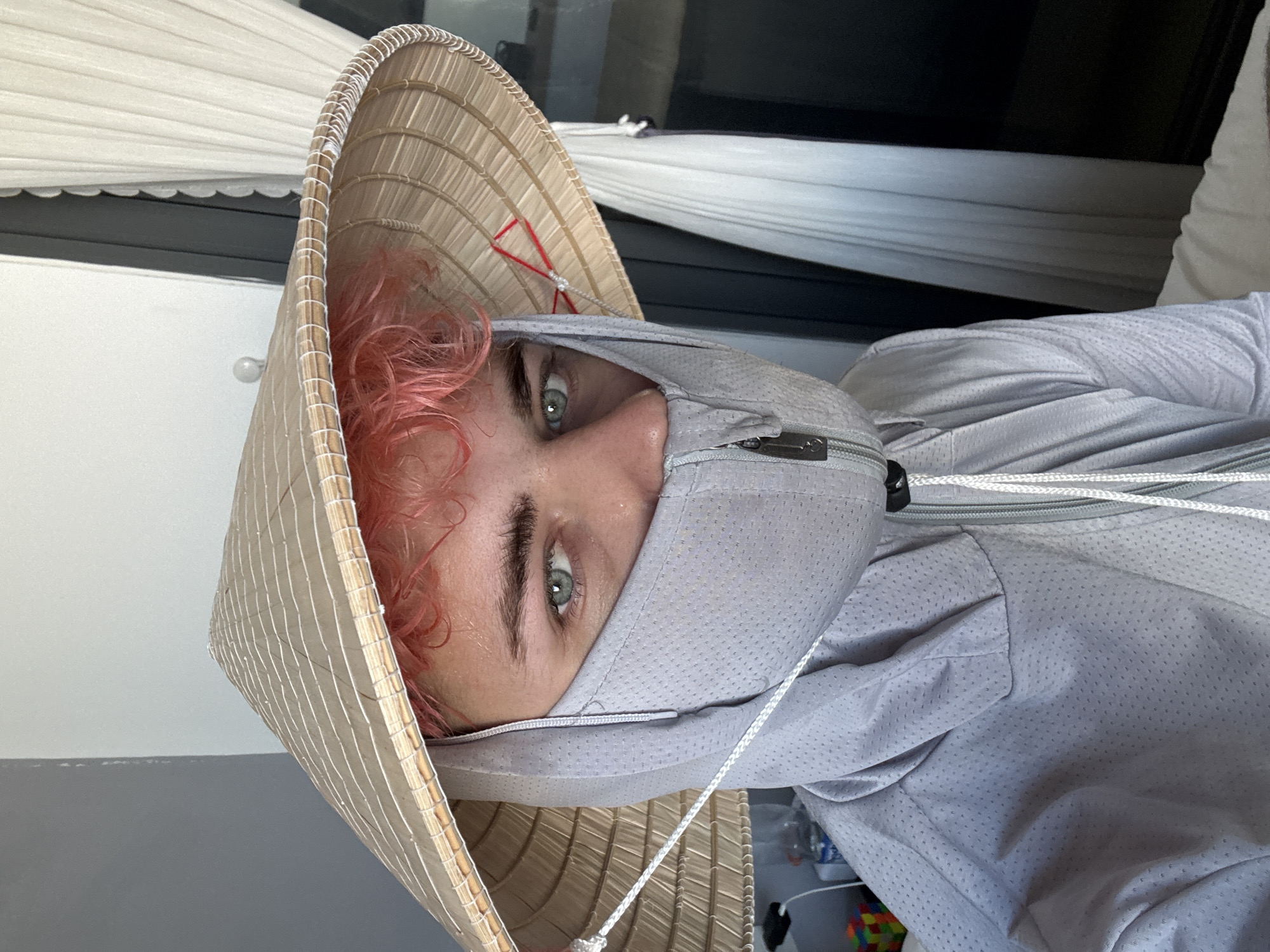 A person with pink hair and blue-grey eyes is wearing a conical Asian hat and a light grey, hooded jacket with a face covering. The jacket is made of a perforated material. A white cord is attached to the hat. The person is indoors near a window with white curtains and blinds. The location is not discernible as Đà Nẵng, Vietnam, based on the image.