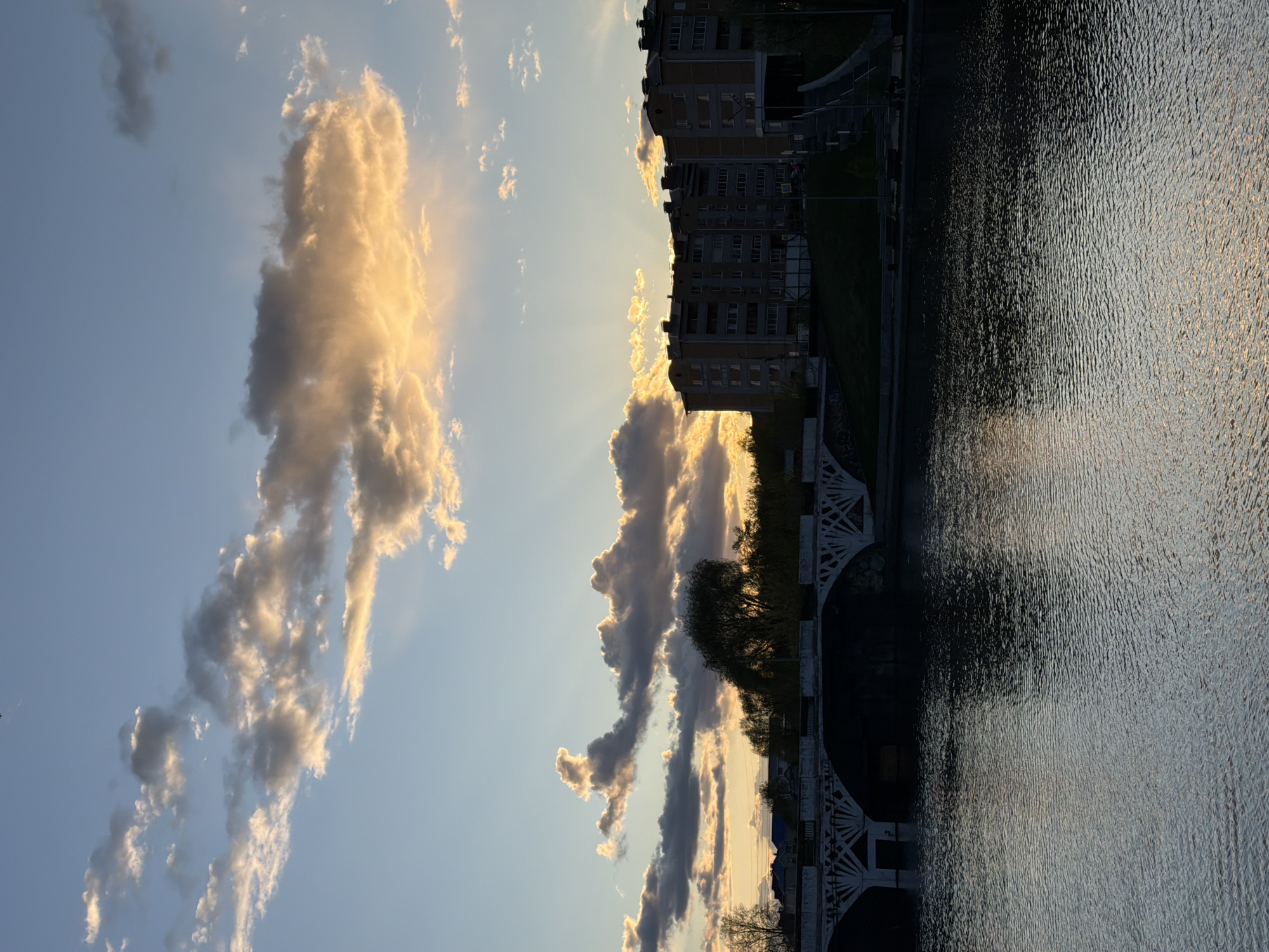 The image shows a waterfront view at sunset in Votkinsk, Russia. Buildings line the edge of the water, which reflects the light of the setting sun. A bridge with decorative white arches spans a section of the waterfront. In the sky, clouds are illuminated with golden light.