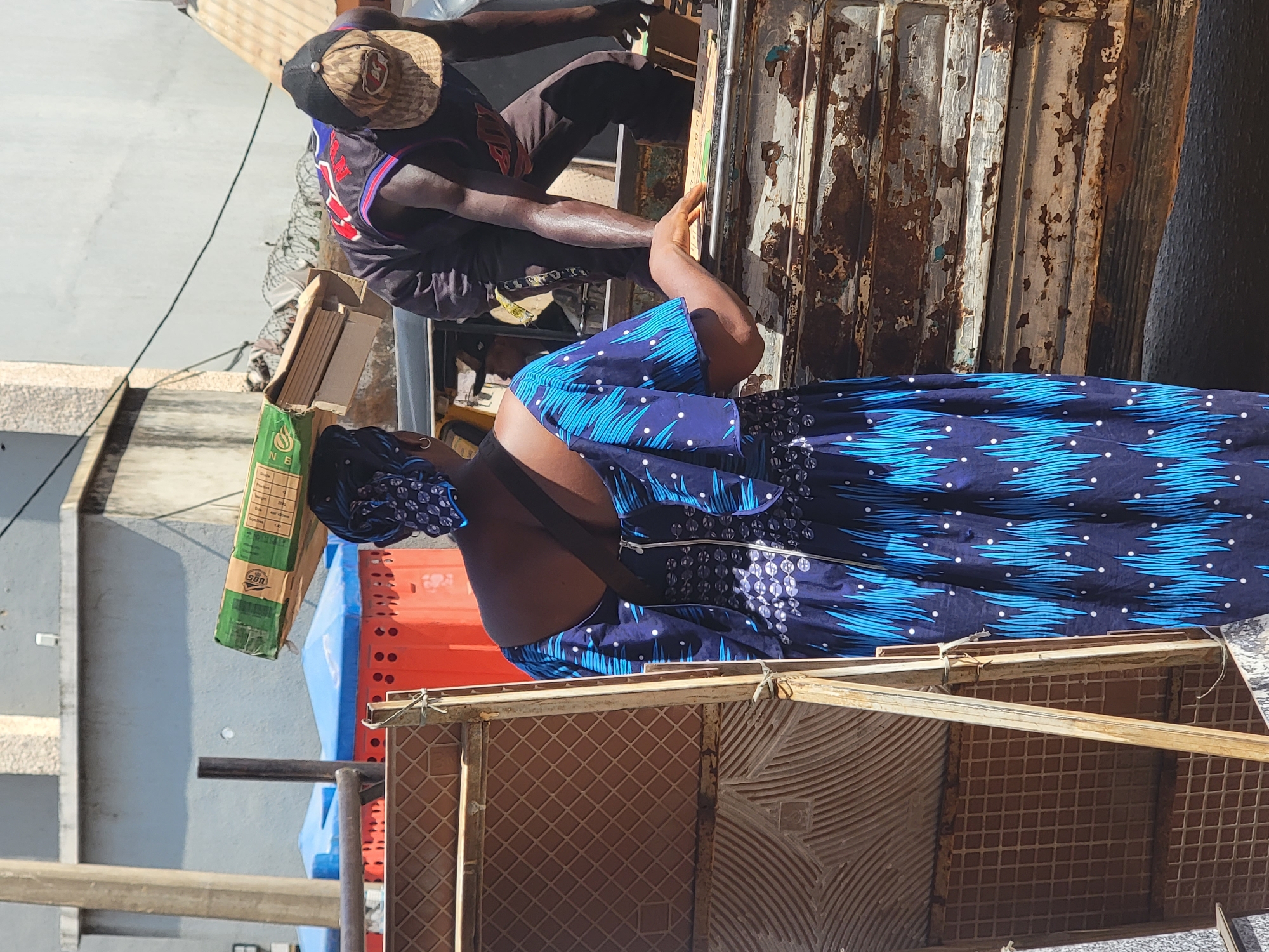 The photo captures a scene in Maiburiji, Nigeria, featuring two individuals alongside construction or transport-related objects. A person wearing a hat and sports jersey is positioned atop a rusty, corrugated metal structure. The person seems to be handling or securing objects. Below, another person wearing a patterned blue dress is interacting with stacked orange crates and a framed object covered with brown tiles. A green package, along with boxes containing wooden planks, are visible. The setting appears to be a bustling outdoor location within the urban environment.