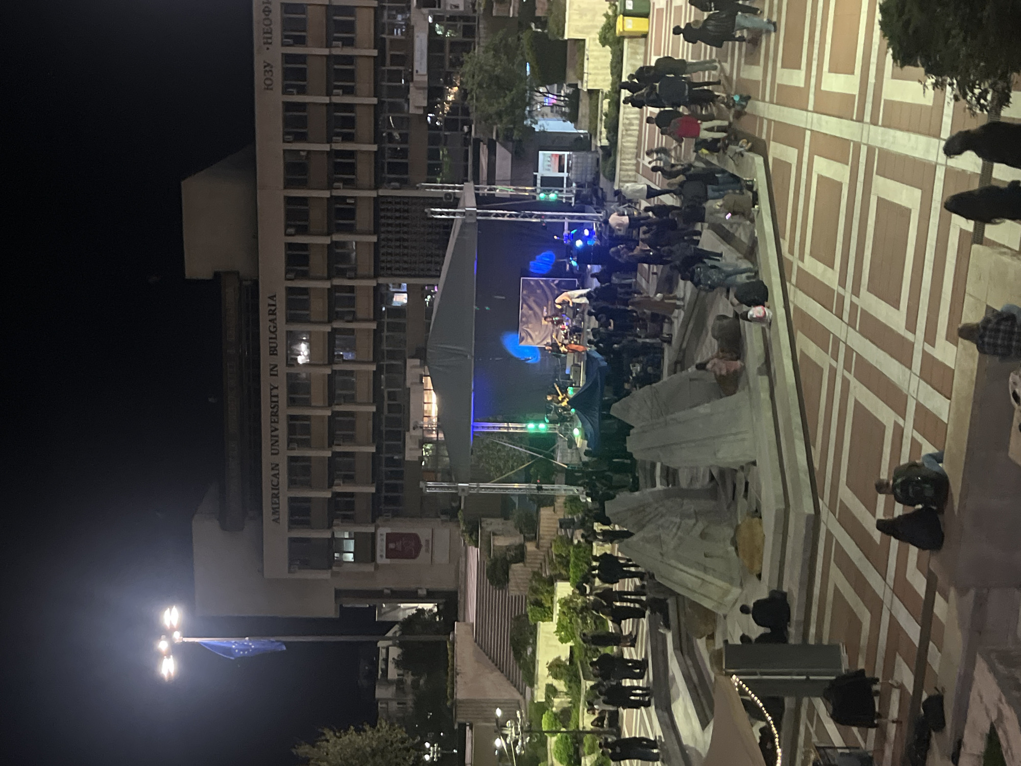 A nighttime shot of the American University in Bulgaria. A stage has been set up in the courtyard in front of the university, and a band is performing, with several people watching. A fountain is in the foreground. Buildings surround the area and the courtyard is lighted.