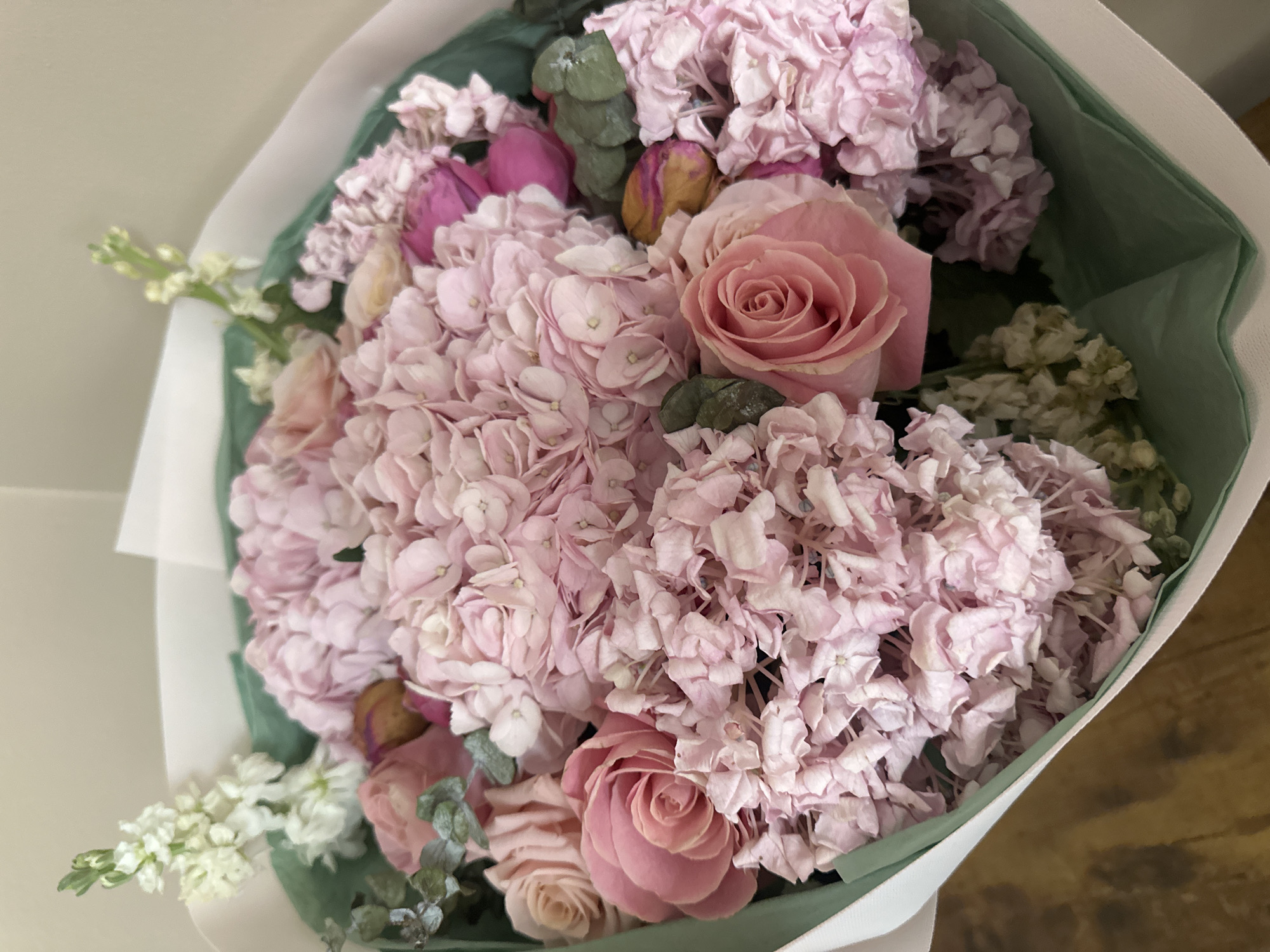 A bouquet of pink and white flowers is displayed in a white paper wrapping. The bouquet is arranged in a close-up, overhead shot and is composed of various flowers, including pink hydrangeas, pink roses, and white stock flowers. Some of the flowers have green foliage. The background is a plain, light-colored wall and a wooden surface. The image was taken in Schaumburg, United States of America.