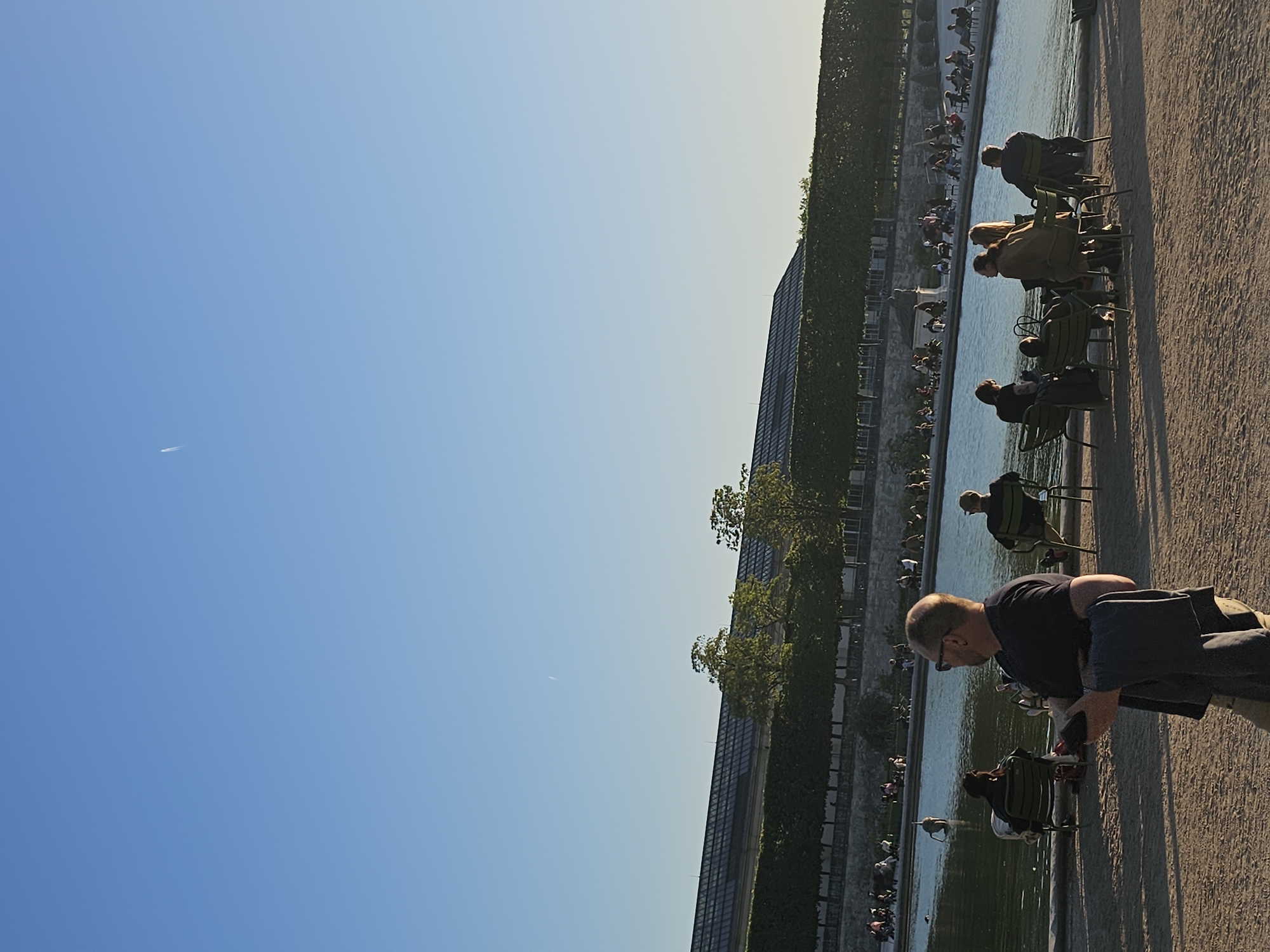 The image captures an outdoor scene in a public park or garden, consistent with locations found in Paris, France. A wide, sandy or gravel pathway runs along the right side of the frame, bordering a large, calm body of water that extends horizontally across the middle of the image.
Numerous people are visible, primarily seated in characteristic green metal chairs along the edge of the pathway, facing the water. In the foreground, a man with short, light-colored hair and glasses, wearing a dark t-shirt, is seated in a green chair, looking down at an object held in his hands. Behind him, other individuals and small groups are also seated, enjoying the serene atmosphere. Many of them are seen in silhouette against the brighter water.
Across the water, more people are scattered, some seated and others walking along another distant bank. A long, modern-looking building, possibly with a glass facade or roof, stretches across the background, partially covered by lush, green foliage and trees.
The sky, occupying a significant portion of the left side of the image, is clear and blue, exhibiting a gradient from lighter near the horizon to a deeper blue overhead. A faint white contrail from an aircraft is visible in the upper left corner of the sky. The lighting suggests late afternoon or early evening, with long shadows and a warm glow on the pathway, contributing to a tranquil and relaxed ambiance. No visible text is discernible in the image.