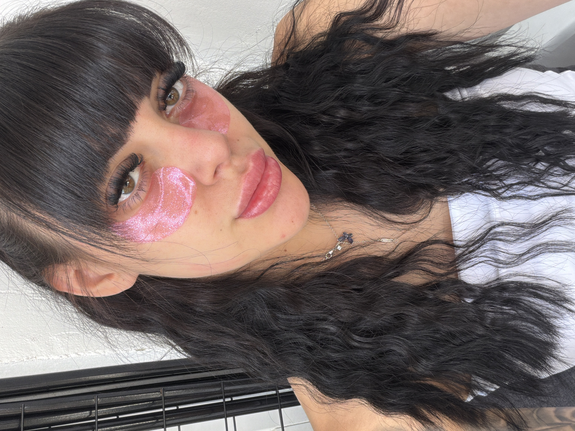 The image is a close-up, head-and-shoulders shot of a young woman with long, dark, wavy hair and bangs. She has on pink under-eye patches, dramatic false eyelashes, and glossy pink lipstick. She is wearing a white top and a delicate silver necklace with a dark pendant. The background is a plain white wall and a black metal fence. The image does not provide any specific information about the location within the city of Plovdiv, Bulgaria.