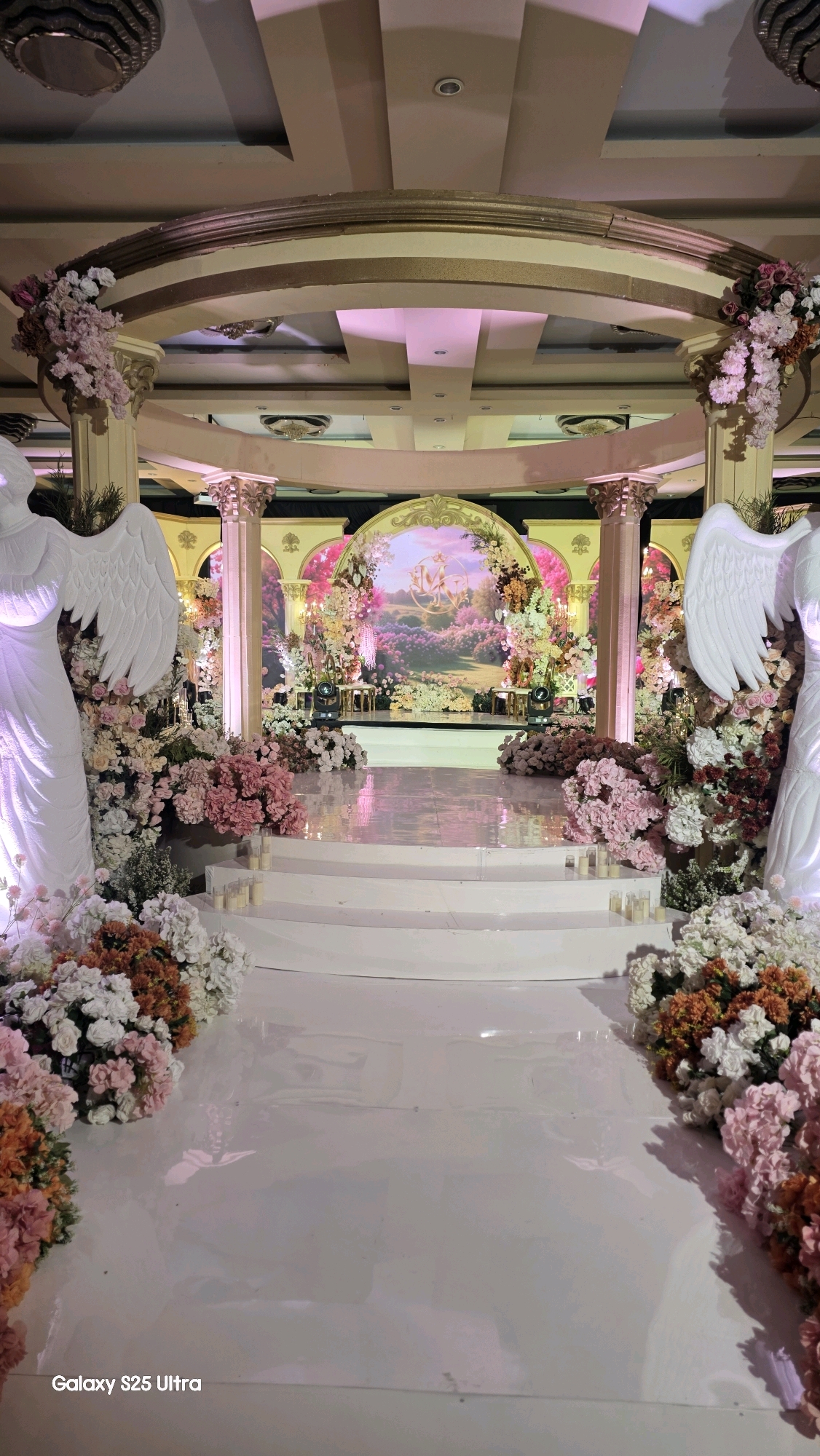 The image depicts a grand and ornate wedding venue, likely in Kecamatan Pati, Indonesia, decorated for a ceremony. In the foreground, a white, reflective aisle leads towards a raised stage. Flanking the aisle are two large, white angelic statues with outstretched wings. The area is extensively adorned with floral arrangements featuring a profusion of pink, white, and coral-colored flowers, interspersed with greenery. Small, lit candles are placed along the sides of the aisle and on the steps leading to the stage.
The stage itself is a raised, multi-tiered platform with a glossy white surface that reflects the surrounding decorations and lighting. Behind the stage, a large, arched backdrop features a painted landscape scene with a castle or manor, surrounded by lush greenery and flowers. The backdrop is framed by decorative golden arches and further enhanced by more floral displays and illuminated columns. The overall lighting is soft and ambient, with pink and purple hues cast from unseen sources, contributing to a romantic and elegant atmosphere. The ceiling features intricate architectural details and elaborate light fixtures. The "Galaxy S25 Ultra" watermark is visible in the bottom left corner of the image. There are no people visible in the scene.