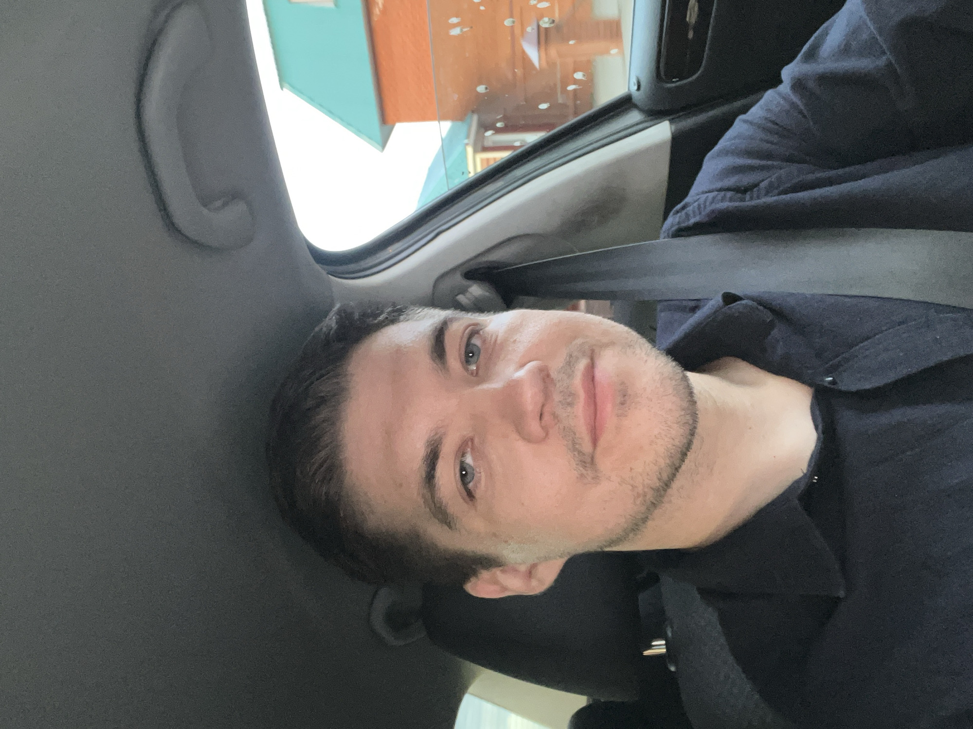 The image shows a man with light skin, short dark hair, and blue eyes wearing a dark button-down shirt and a seatbelt. He is inside a car. Through the car window, the blurred exterior suggests a building with a light green and orange facade, possibly a storefront or a residential building. The presence of water droplets on the window suggests a recent rain or a damp environment. There is no definitive evidence to confirm the location as Kazan, Russia based on this image alone.