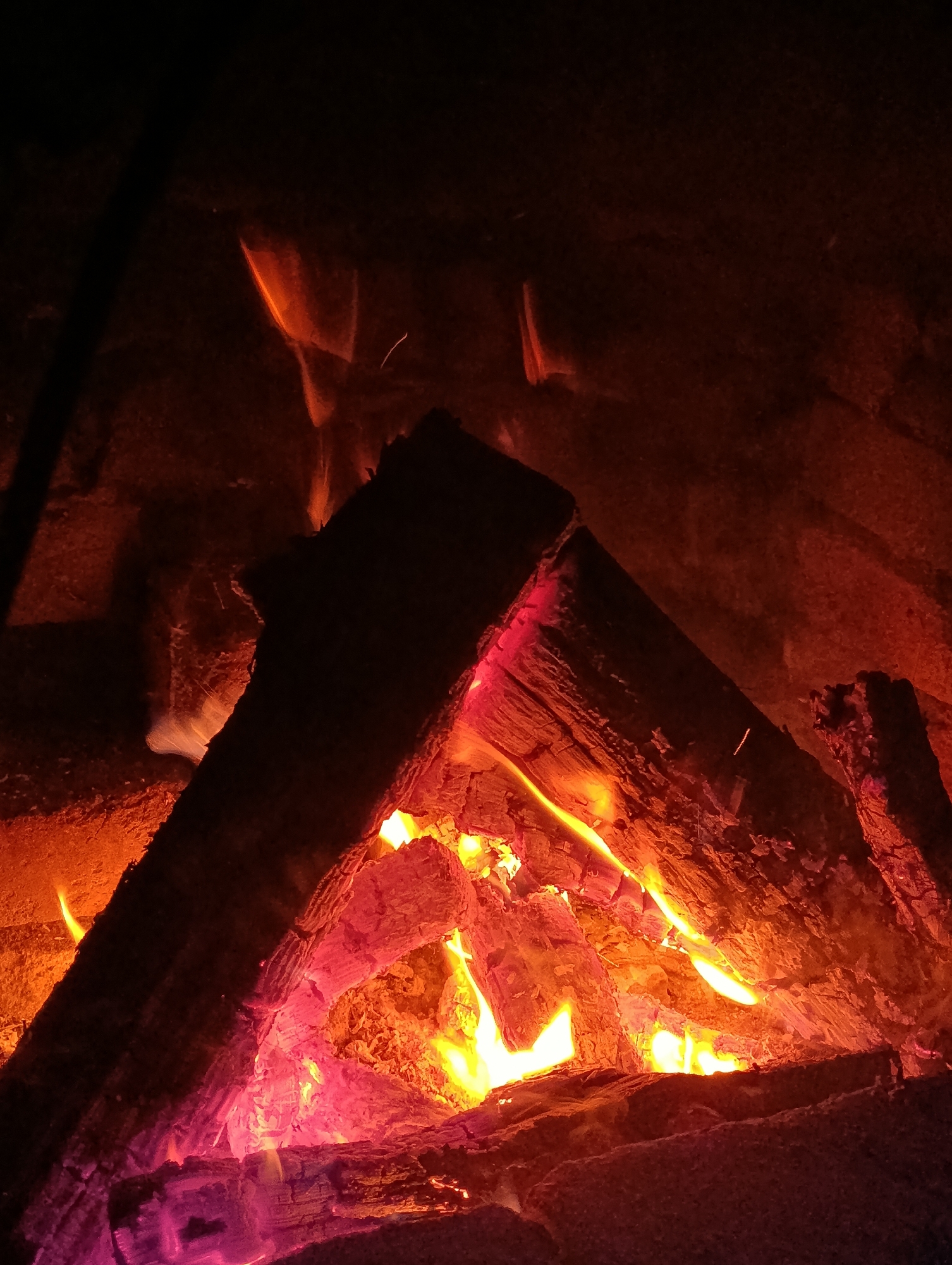 A close-up, low-light photograph captures the intense heat and dynamic nature of a crackling bonfire. The scene is dominated by glowing embers and vibrant flames in shades of orange, yellow, and pink, illuminating charred logs arranged in a triangular formation. The rough texture of the burning wood and the uneven surfaces of what appears to be a stone fireplace are visible. The background is a dark, indistinct mass, suggesting the surrounding environment is also in shadow or obscured by smoke. There are no people present in the image. The location in Данілавіцкі сельскі Савет, Belarus, places this scene in a rural Belarusian setting, likely a backyard or outdoor gathering area where a bonfire is being enjoyed.